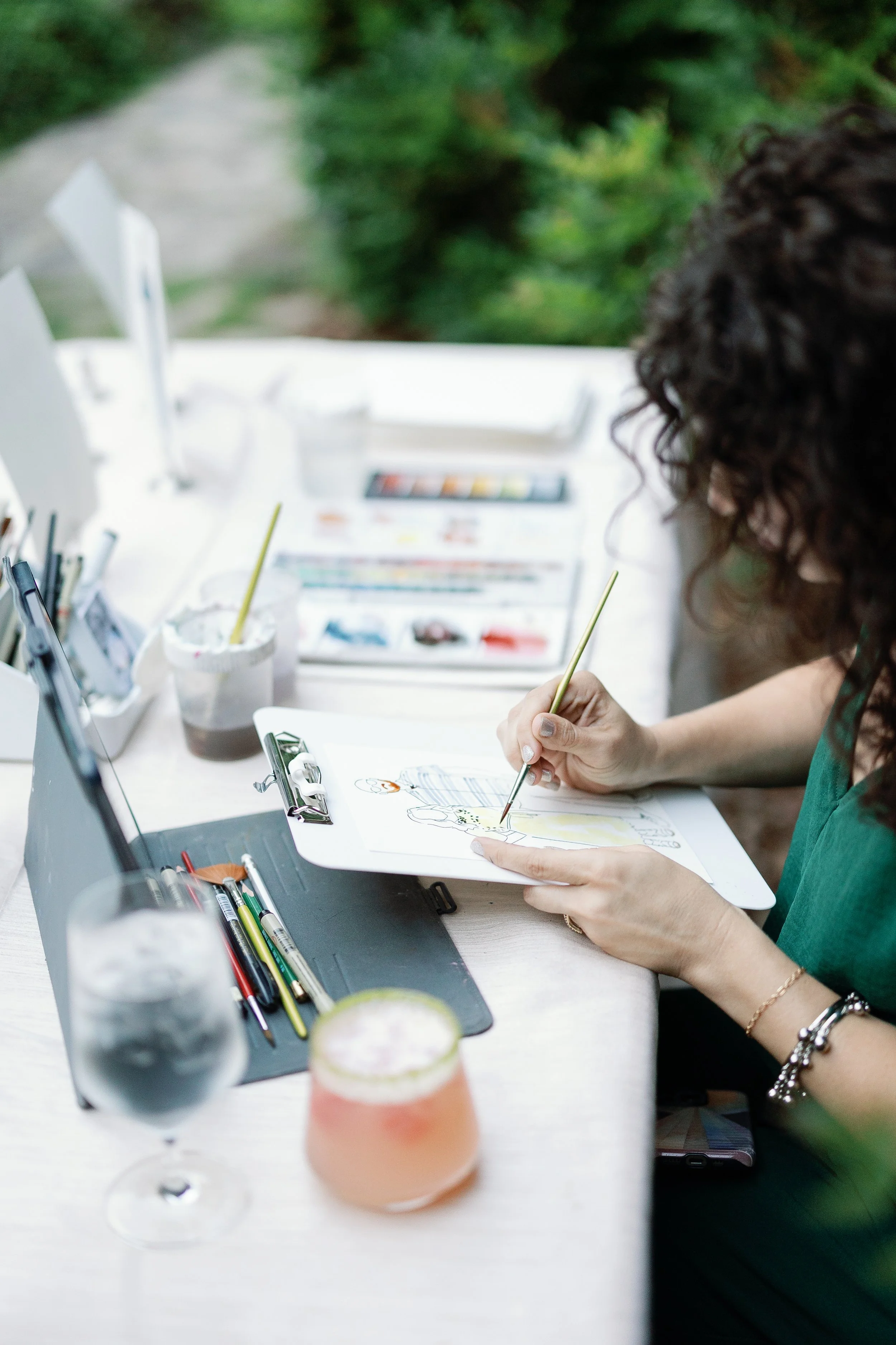 Holly drawing and coloring on a piece of paper at an outdoor table, surrounded by art supplies, beverages, and greenery in the background.