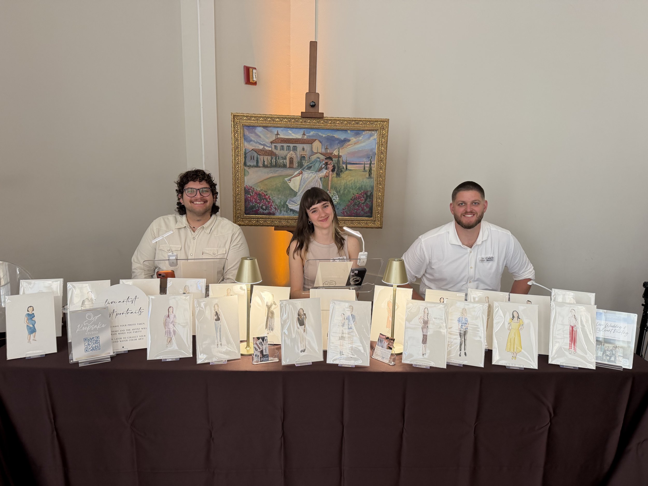 A group of three smiling people sitting behind a table at an art event, with a colorful painting of a woman in a white dress and a large framed art piece in the background. The table displays illustrated art prints and promotional materials.