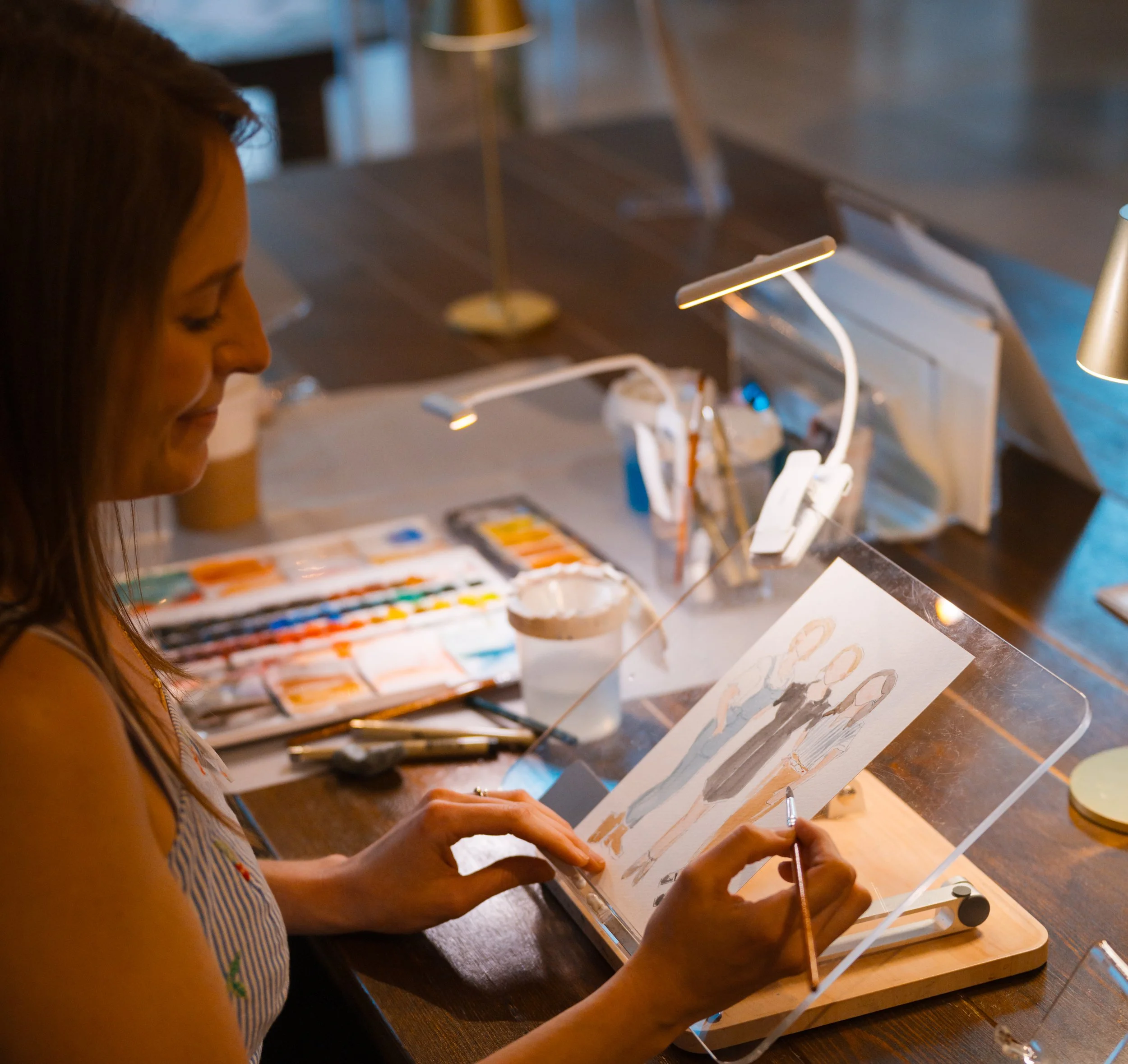 A woman with long brown hair wearing a striped sleeveless top is painting on a canvas in an art studio with warm lighting and various art supplies.