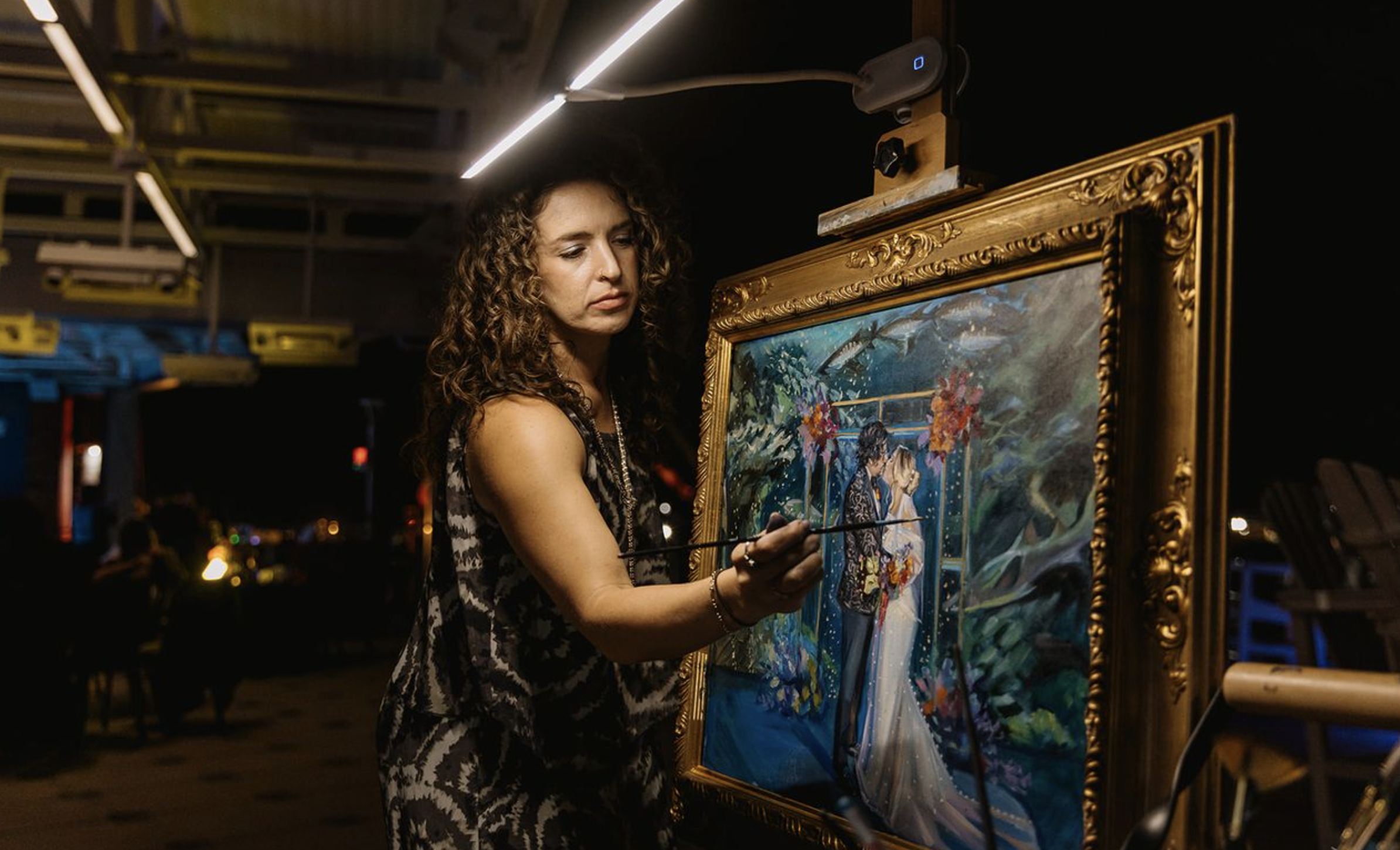 A woman with curly hair painting on a framed canvas at an indoor art event, with a dark background and some blurred lights.