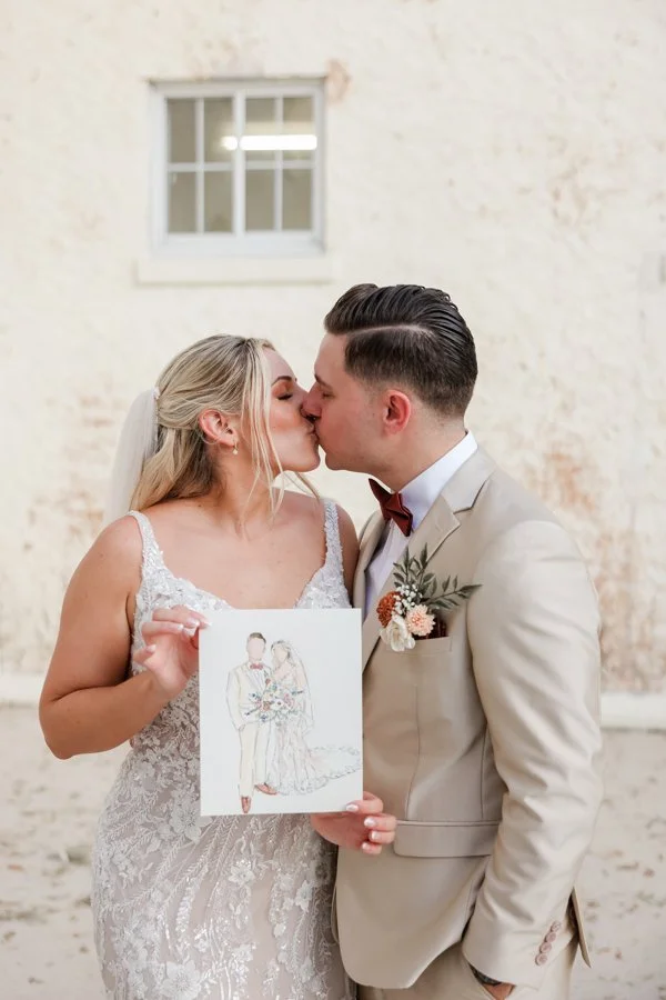 Bride and groom kissing at their wedding, holding a drawing of them in wedding attire, inside a rustic venue with a brick wall and a small window.
