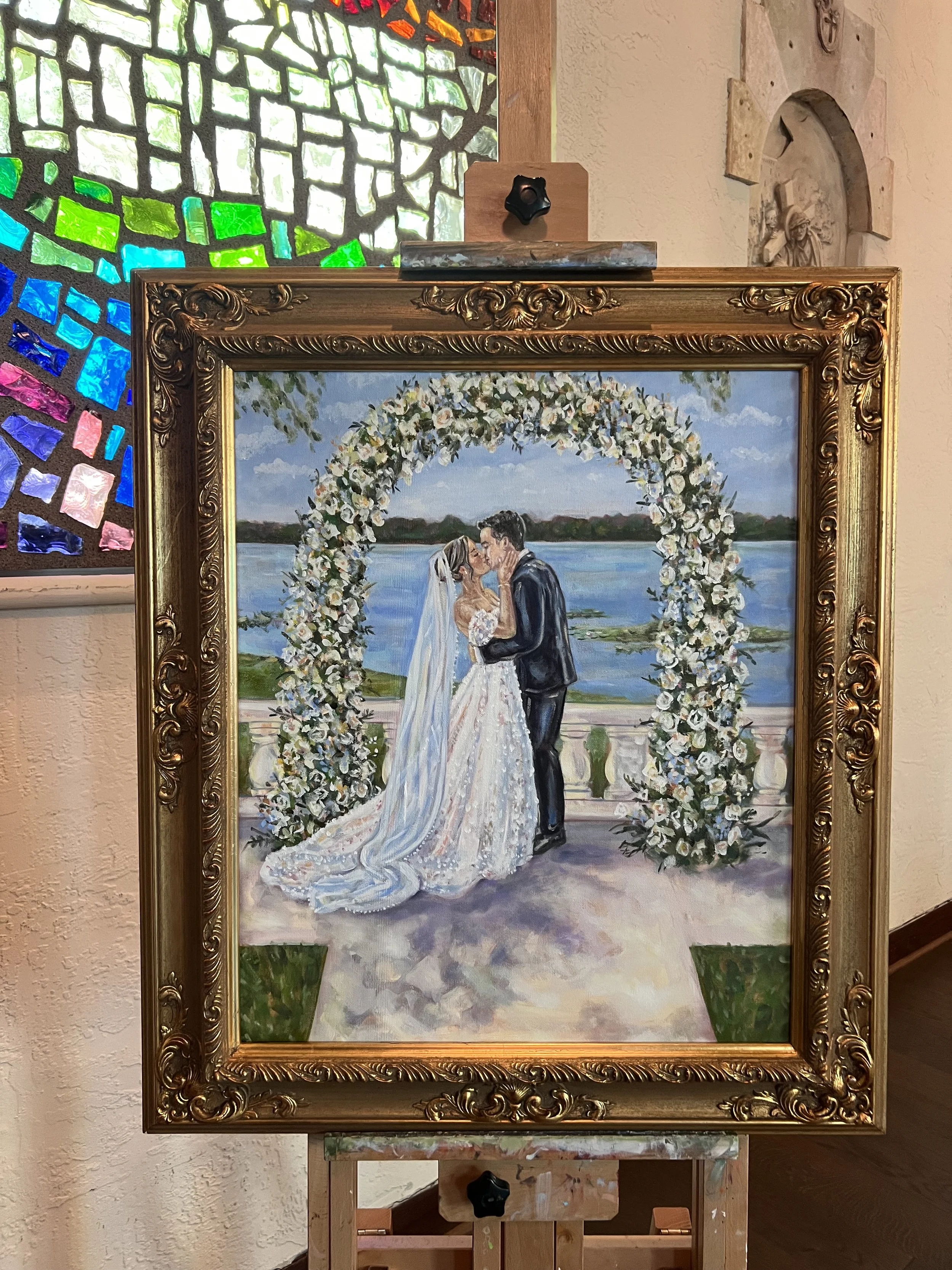 A wedding painting showing a bride and groom kissing under a large floral arch outdoors with water and landscape in the background.