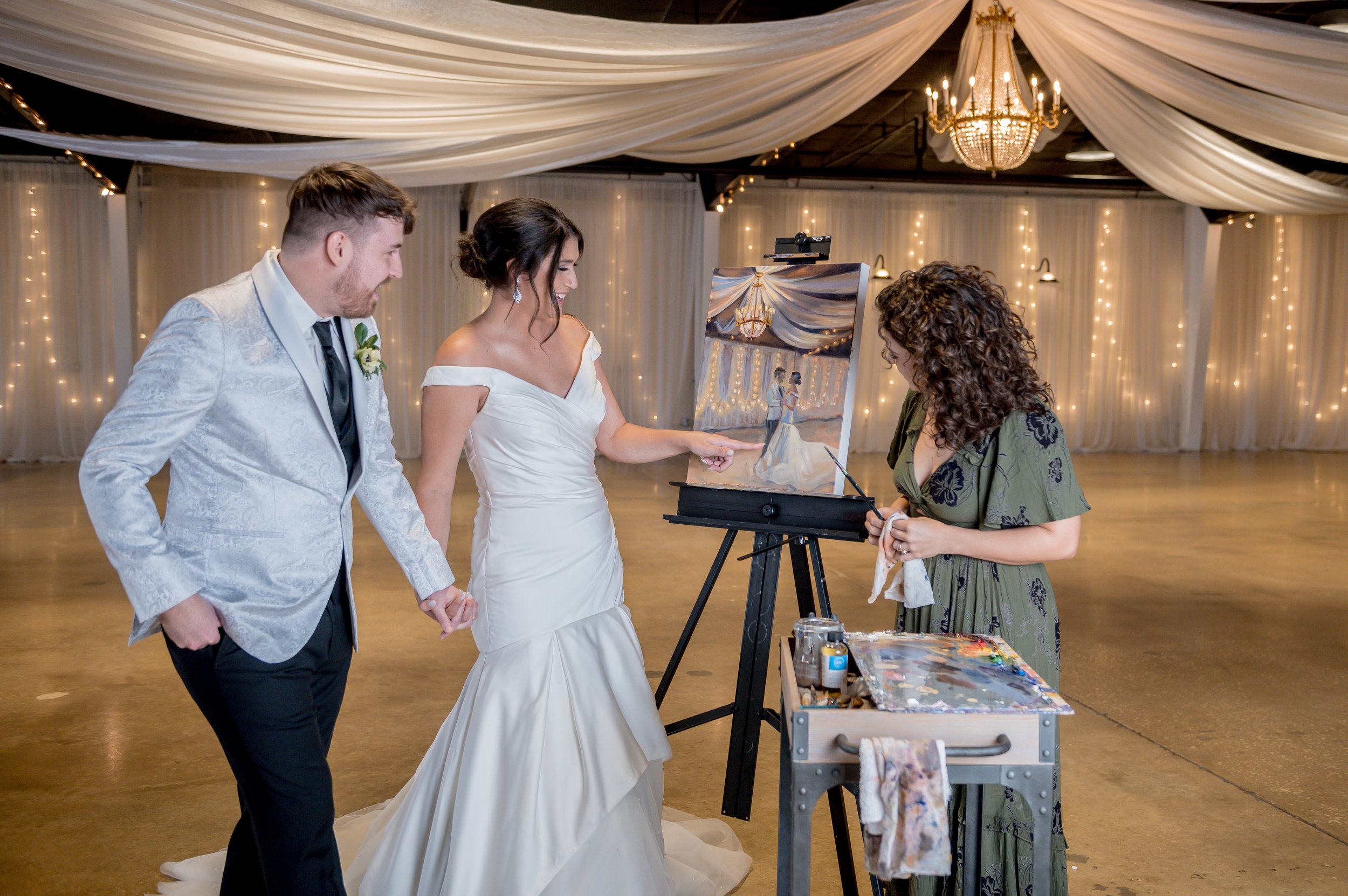 A bride and groom holding hands are watching an artist paint a wedding portrait during their wedding reception. The room is decorated with white drapes and string lights, and a chandelier hangs from the ceiling.