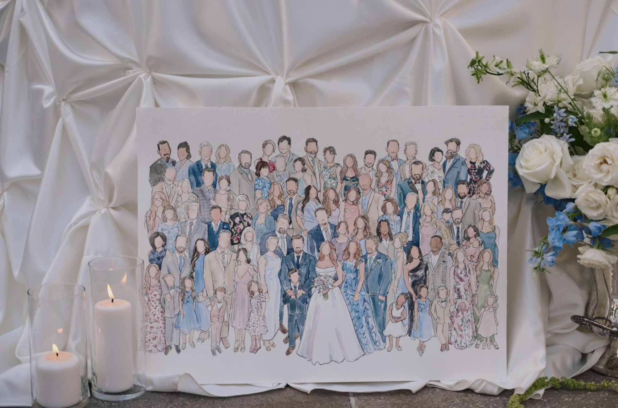 A wedding reception table decorated with white drapery, floral arrangements, candles, and a drawing of a group of people in formal attire.