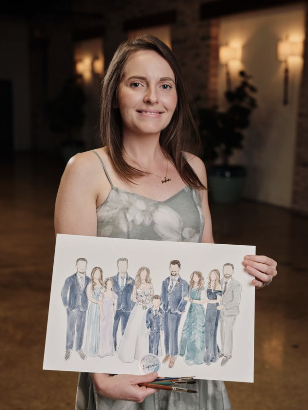 A woman holding a watercolor illustration of a wedding party, including brides and grooms, in an indoor setting with warm lighting and brick walls.