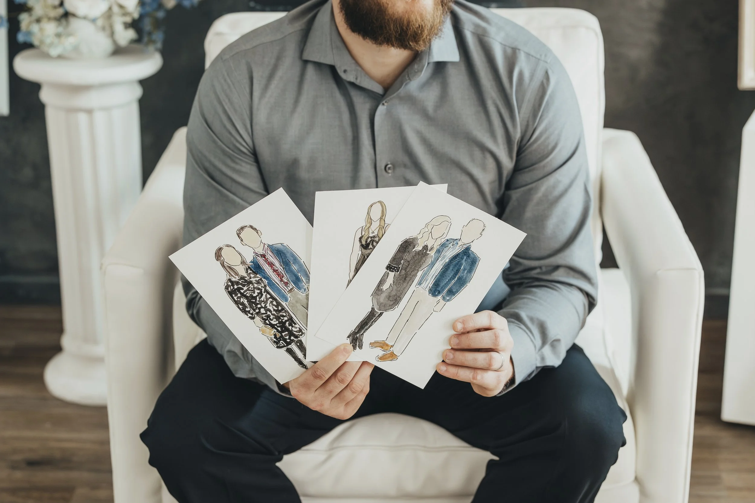 Caleb sitting on a white armchair holding three fashion sketches of models and couples with clothing designs