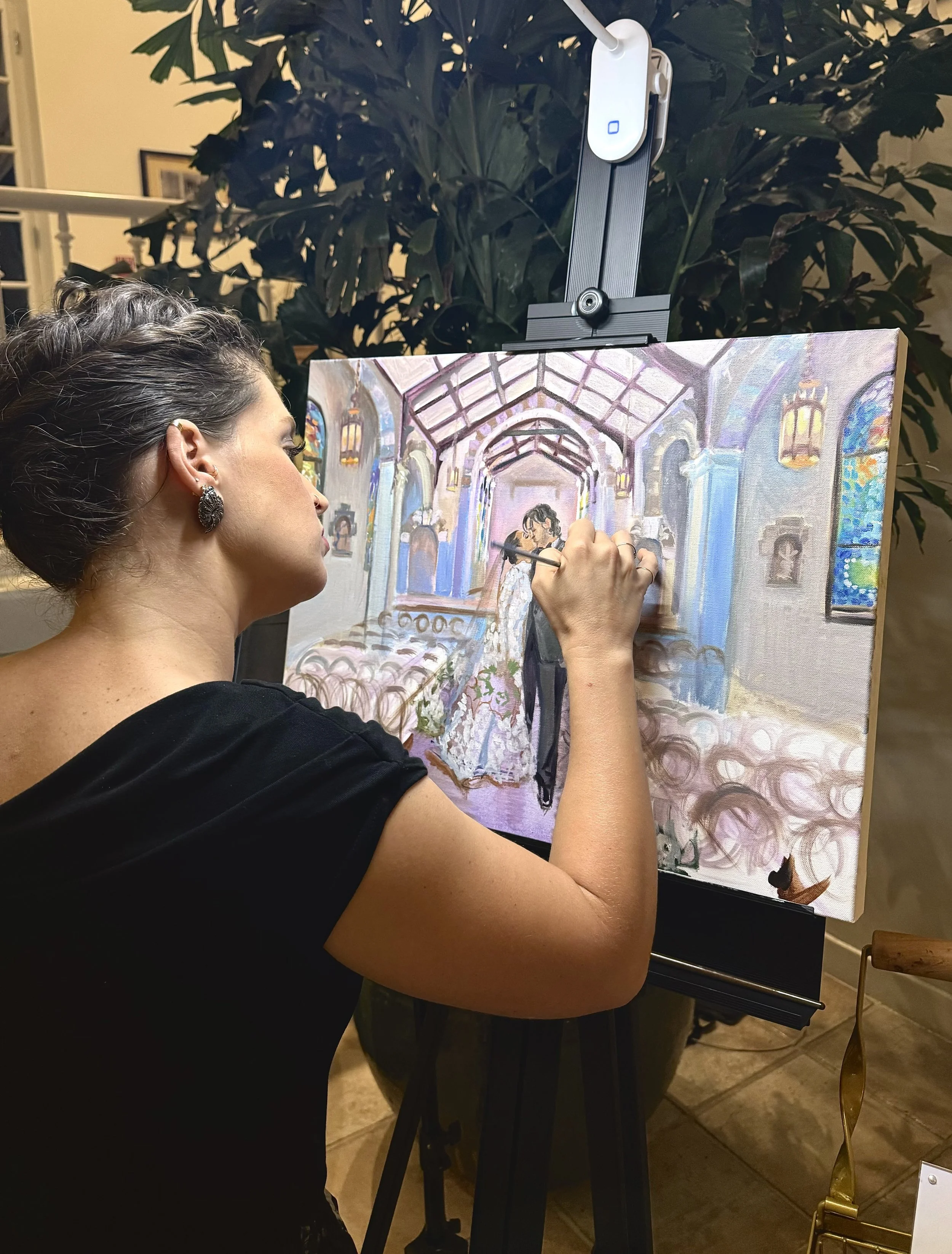 Holly painting a wedding scene on a canvas, with a large indoor plant and warm lighting in the background.