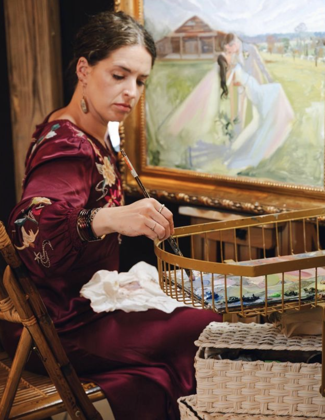A woman with dark hair and earrings, wearing a burgundy floral dress, sitting on a wooden chair, painting on a basket with a paintbrush, with a colorful landscape painting in the background.