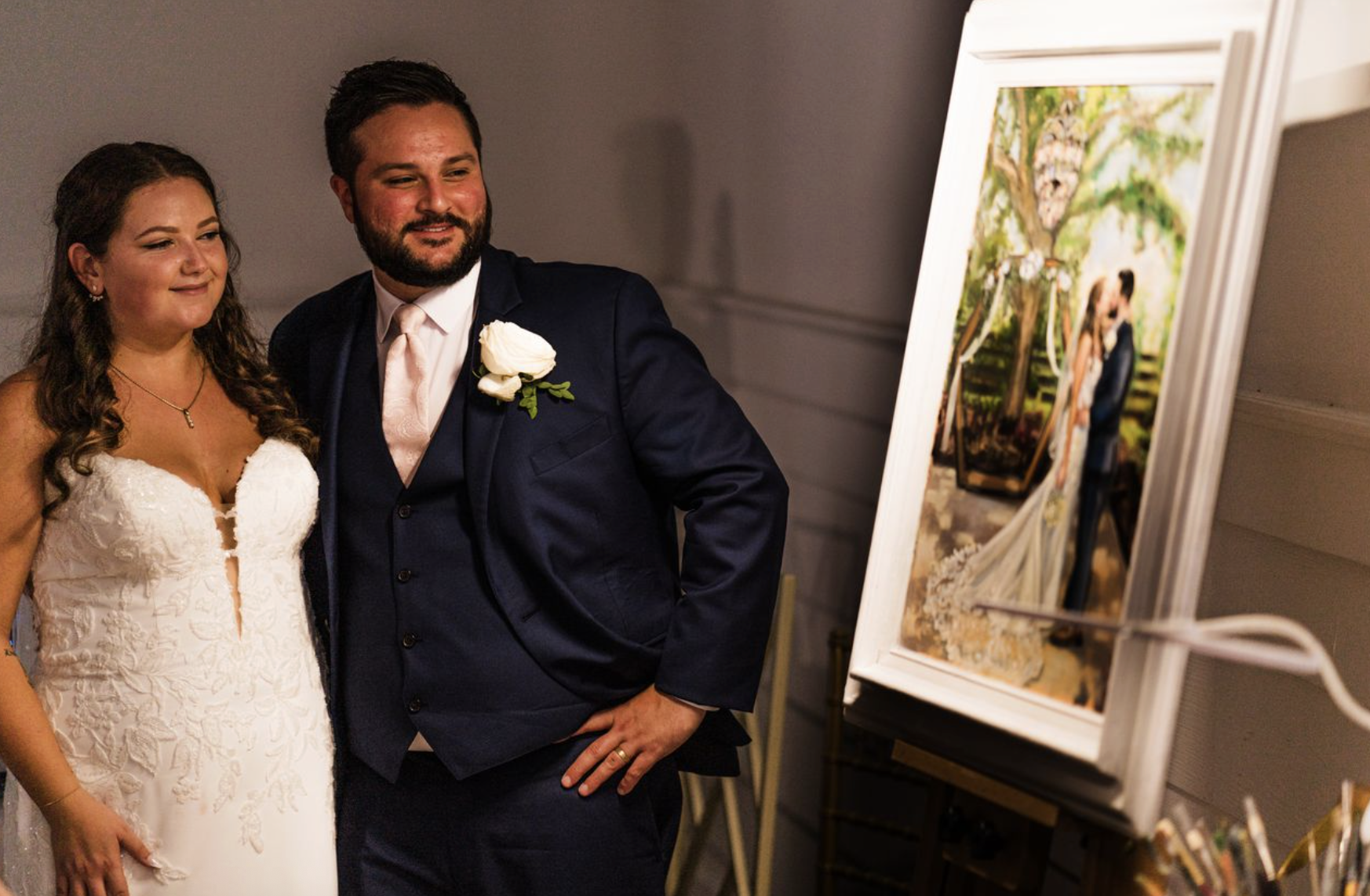 A bride and groom at a wedding reception, standing next to an artwork display. The bride is wearing a white lace wedding gown, and the groom is in a dark suit with a pink tie and white boutonniere. They are smiling and looking at an art piece on a st