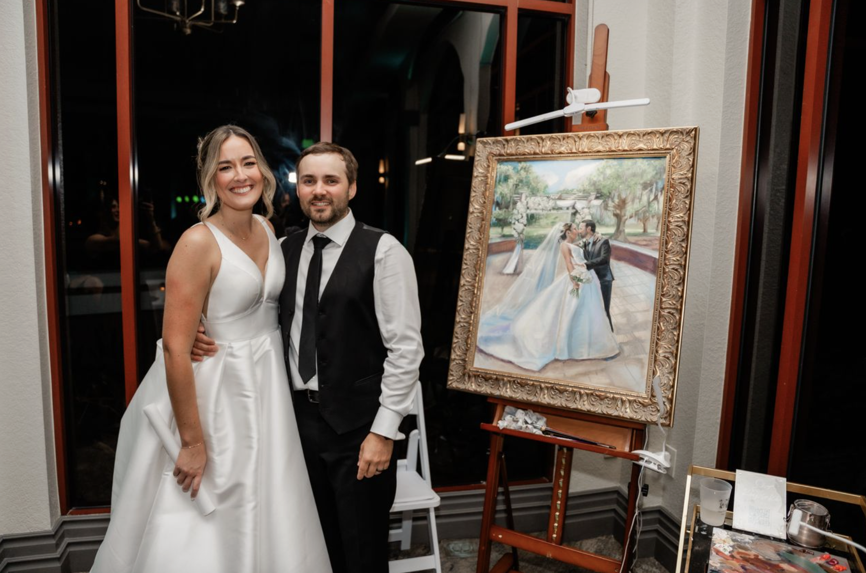 A bride and groom standing together indoors at a wedding reception, smiling at the camera, with a framed wedding portrait painting on an easel beside them.