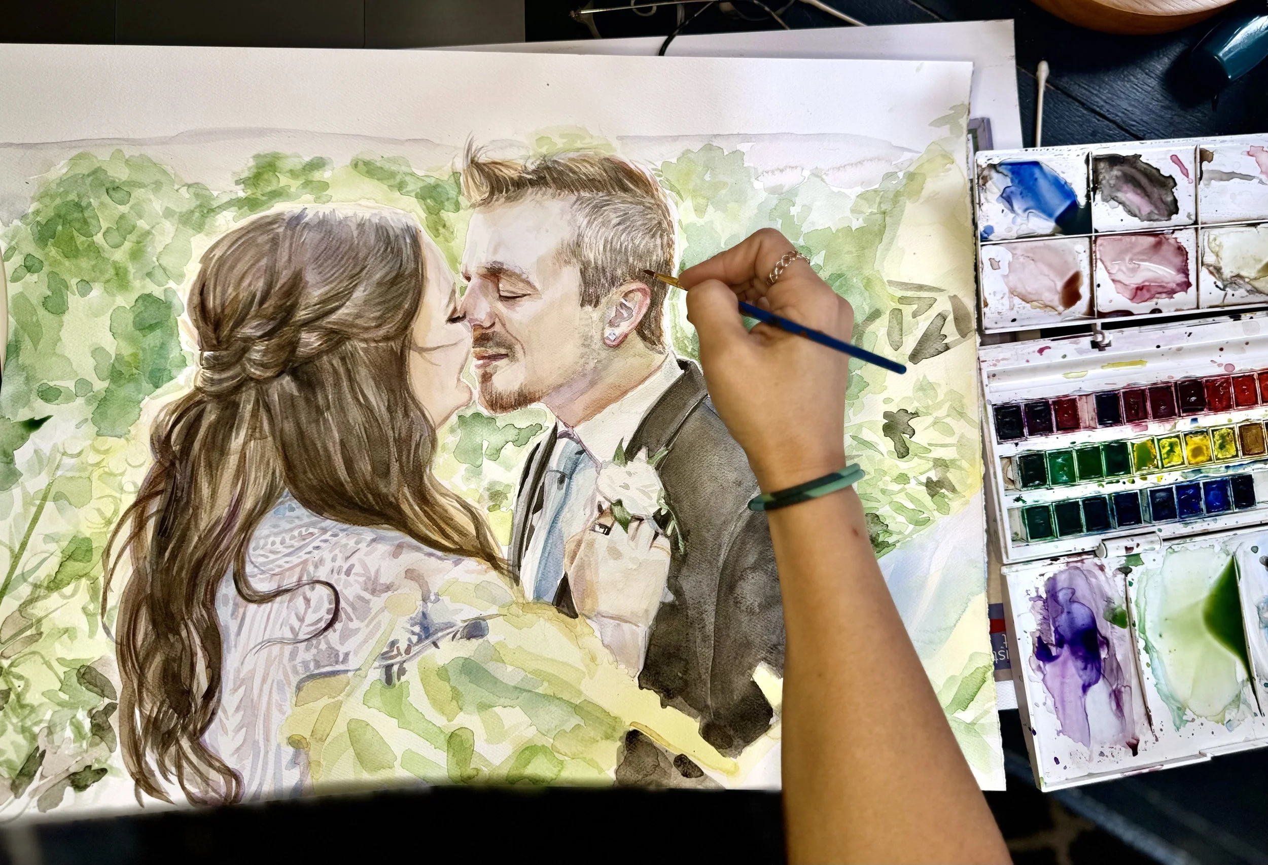 Artist painting a watercolor portrait of a couple on their wedding day, touching noses and smiling, with greenery in the background.
