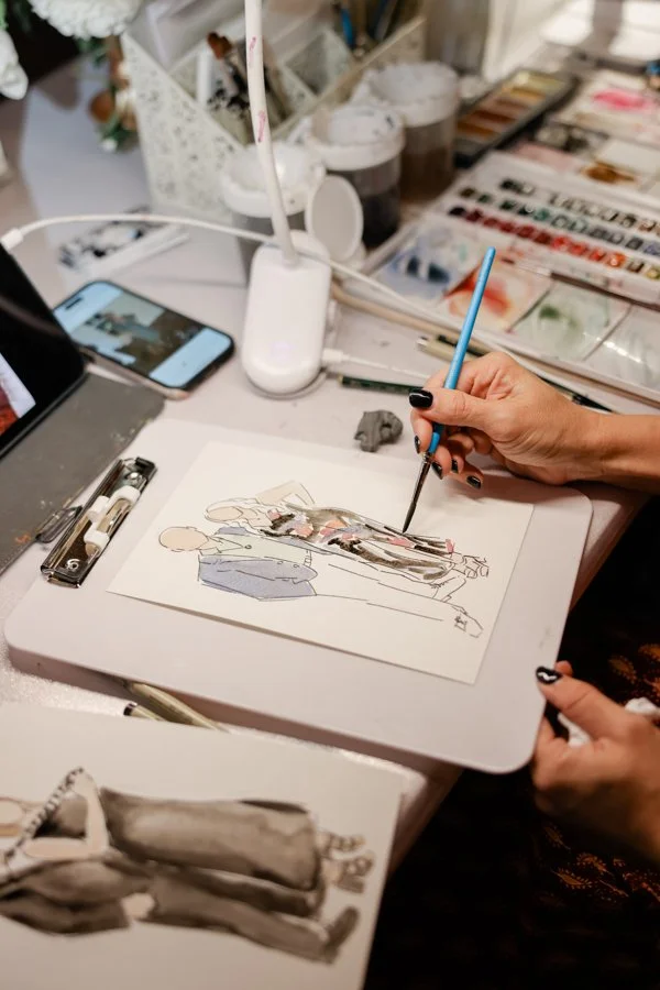 An artist sketches fashion figures on paper at a workspace with art supplies, a phone, and a lamp.