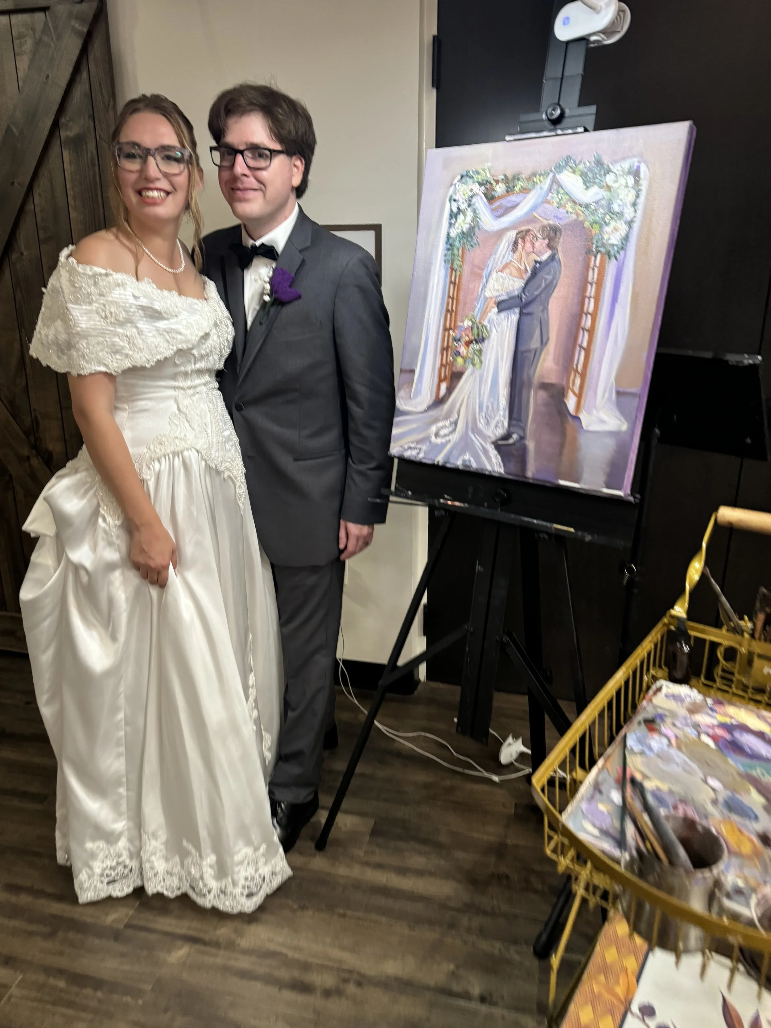 A bride and groom posing next to a wedding portrait painting at their wedding reception. The bride is wearing a white lace wedding dress, and the groom is wearing a grey suit with a black bow tie.