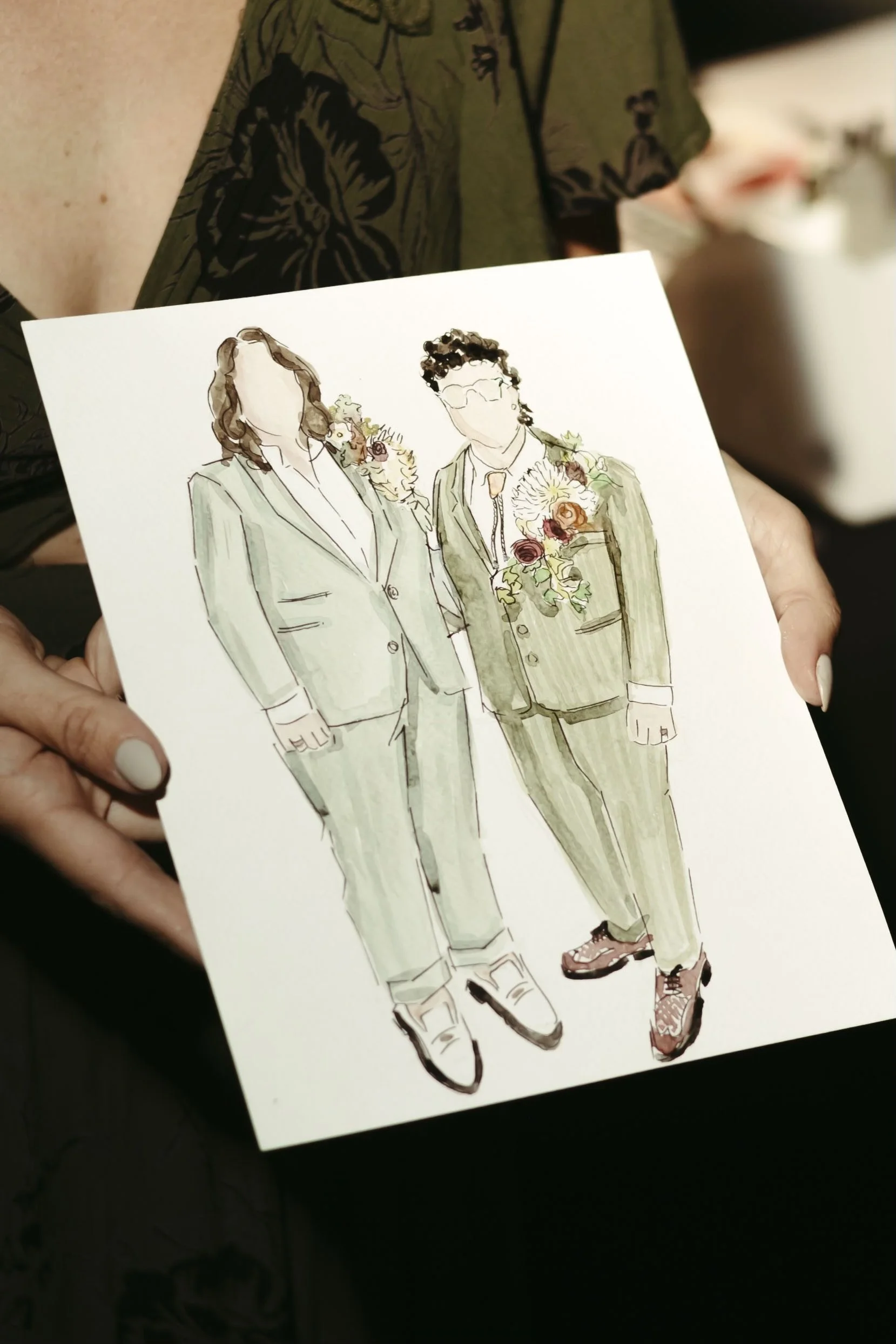 A person holding a watercolor sketch of two women in green suits with floral accents.