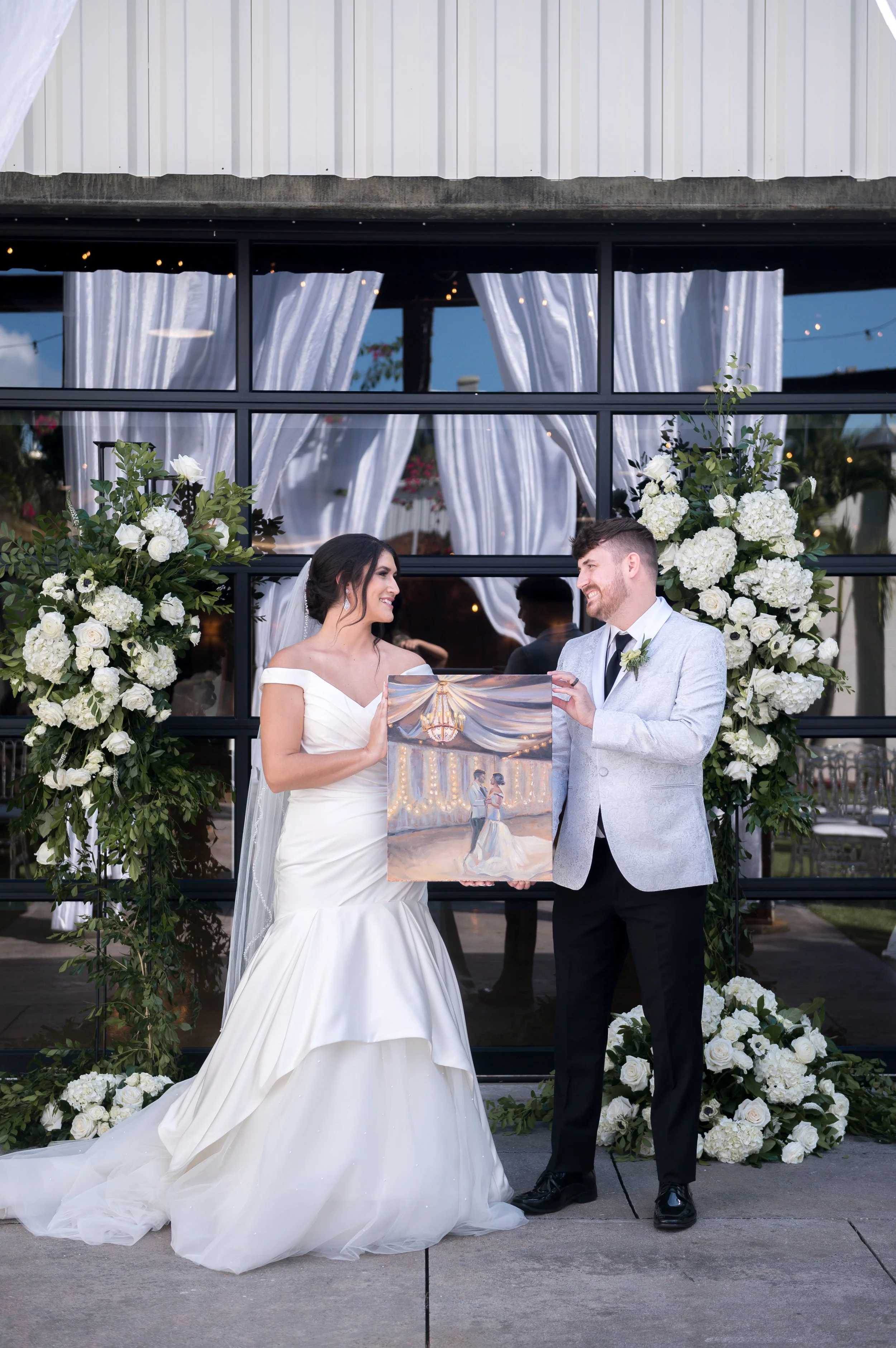 A bride and groom with their painting at their wedding ceremony, standing outdoors in front of decorated floral displays and a glass window with white curtains.