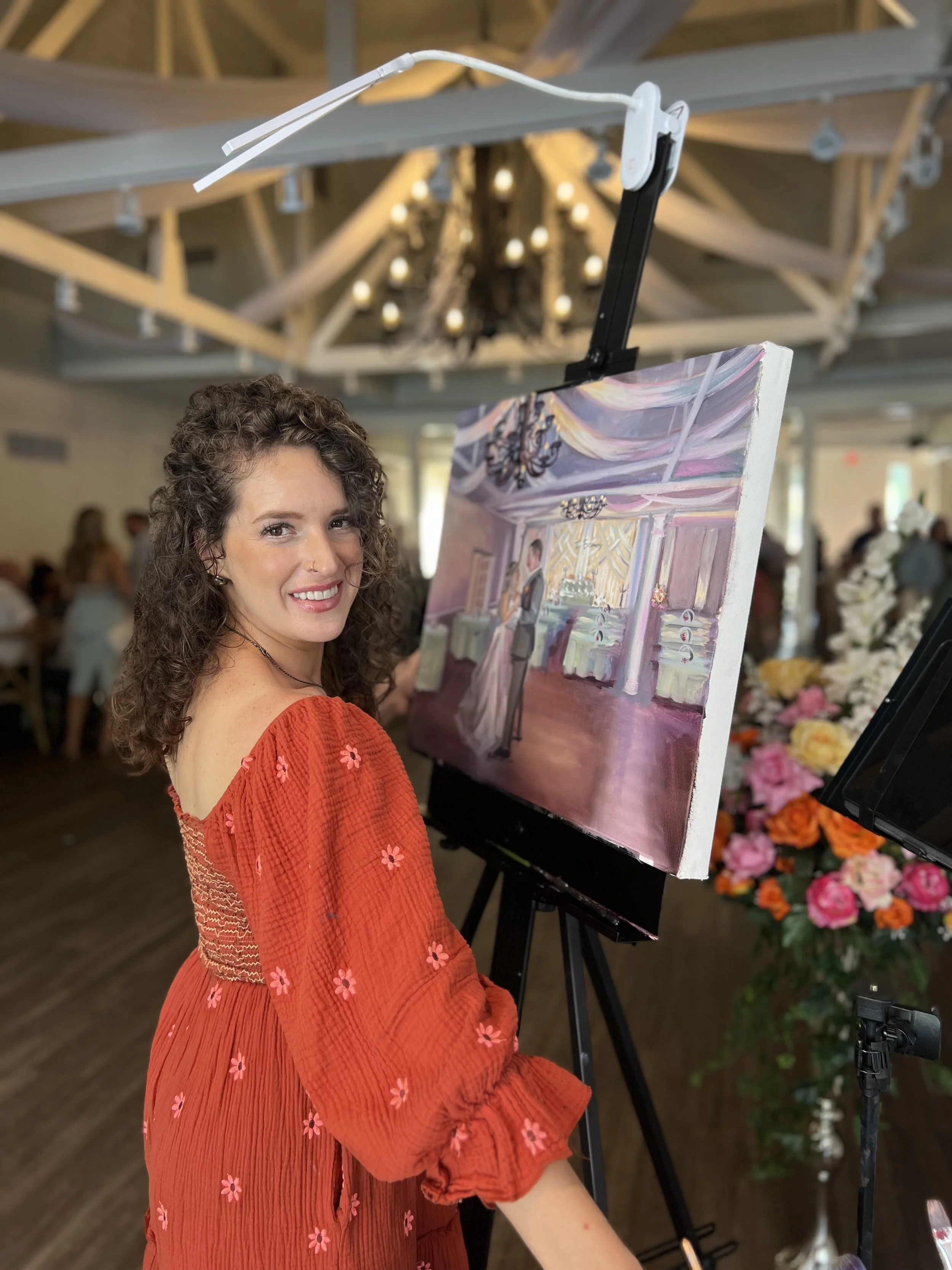 Holly smiling at the camera while standing next to an easel with a colorful painting of an elegant indoor scene, wearing a red dress with pink floral embroidery, in a decorated venue with chandeliers and flowers.