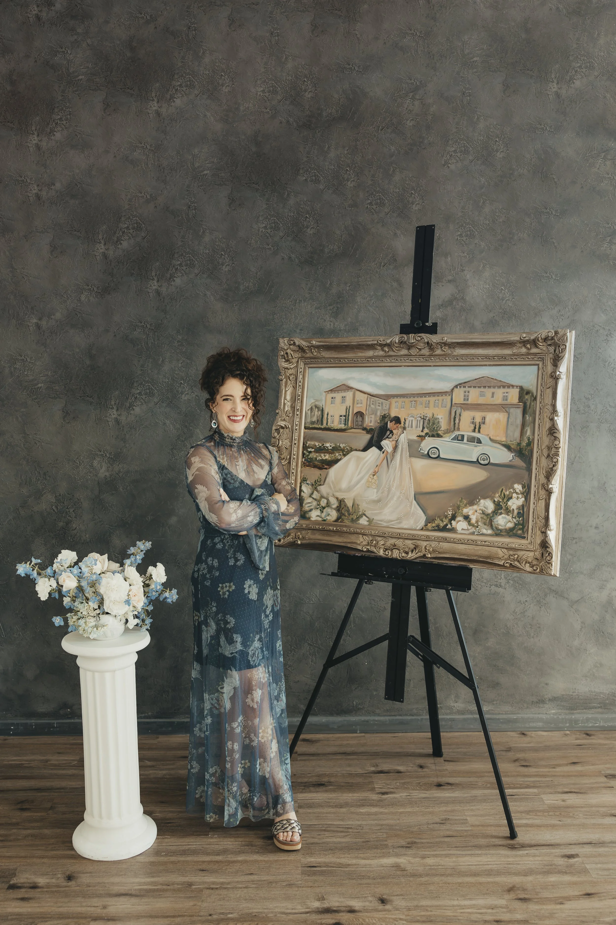 Holly standing next to an easel holding a painting of a couple in a wedding scene, with a flower arrangement on a pedestal nearby, against a dark gray textured wall.