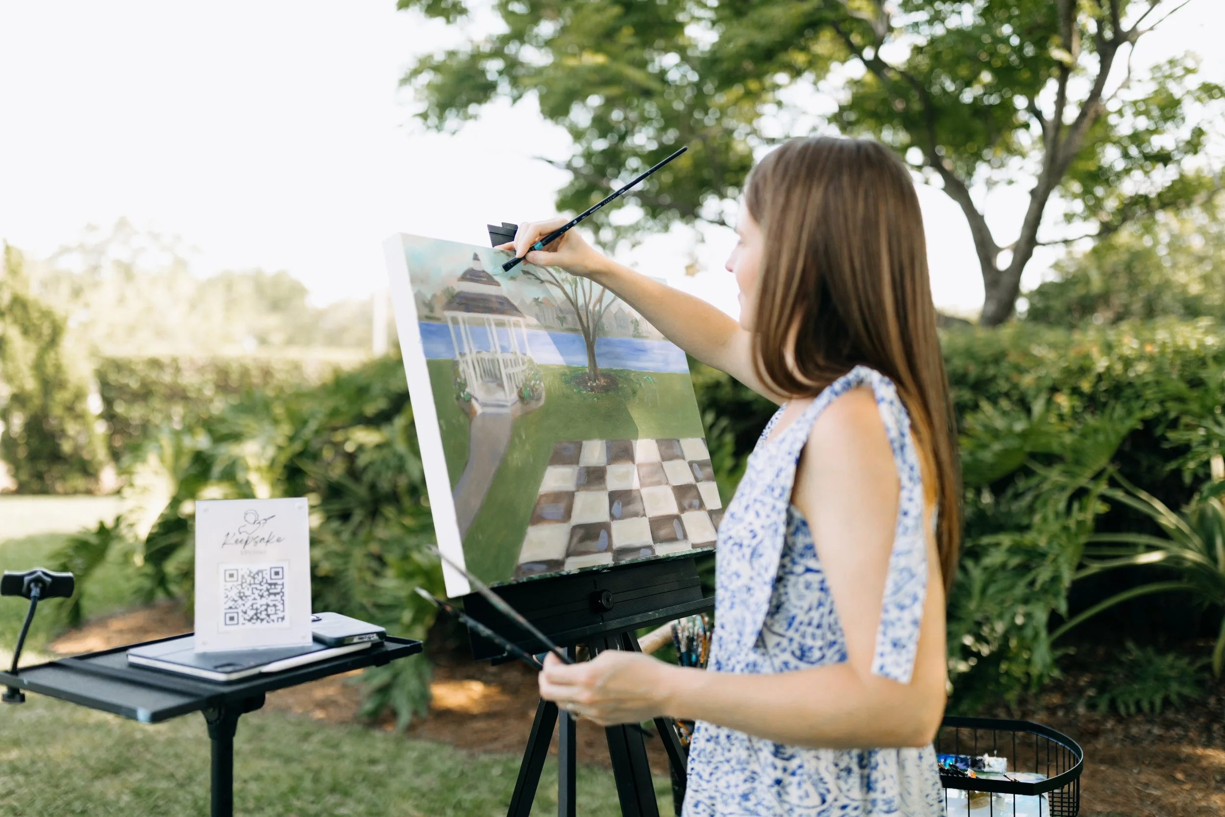 Brittany painting outdoors on a sunny day with green trees and bushes in the background. She is standing at an easel, working on a landscape wedding painting featuring a gazebo, a pathway, a tree, and a body of water.