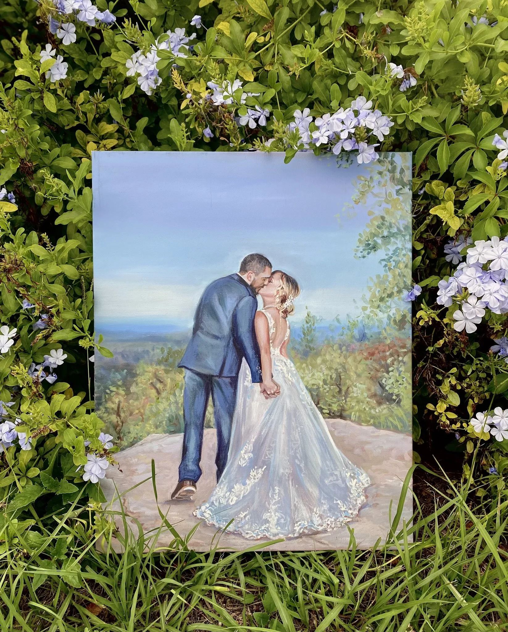 A painted wedding portrait of a couple kissing, with the groom in a blue suit and the bride in a white lace wedding dress, set against a scenic landscape background, surrounded by green foliage and purple flowers.