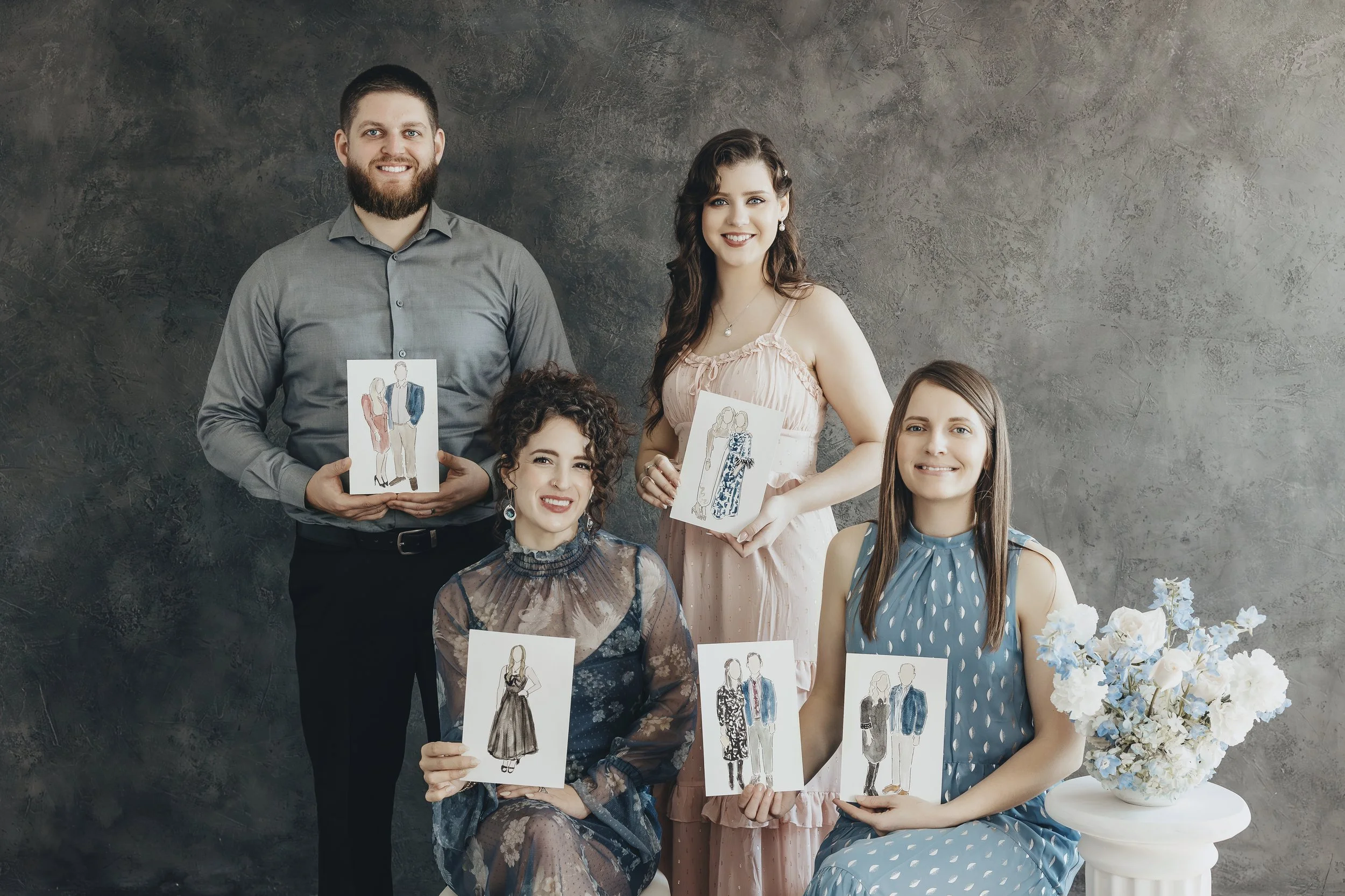 Four artists posing in front of a textured gray wall, each holding a fashion illustration of different outfits, with a white flower arrangement on a small table nearby.