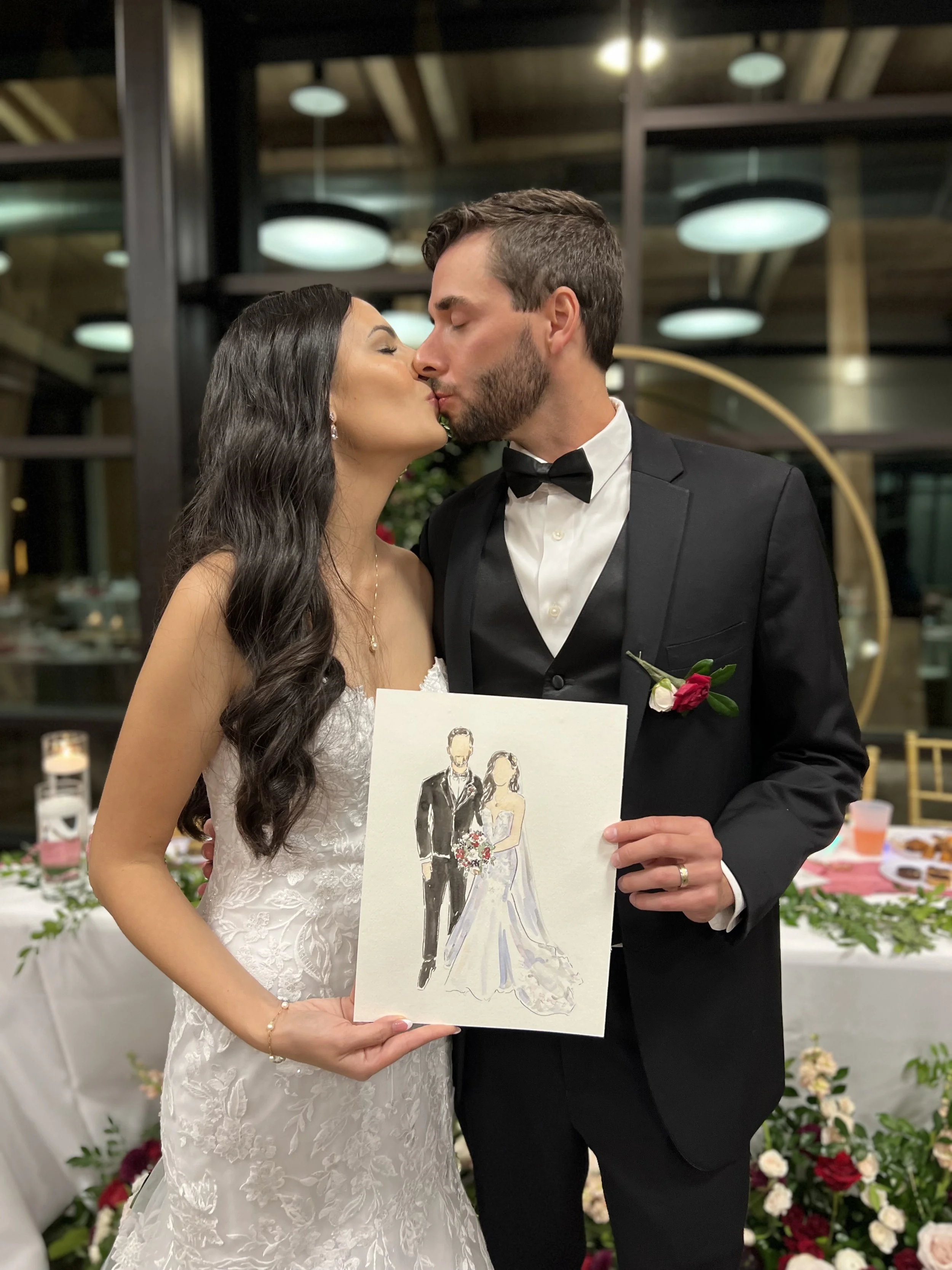 A bride and groom kissing at their wedding reception, holding a watercolor portrait of themselves.