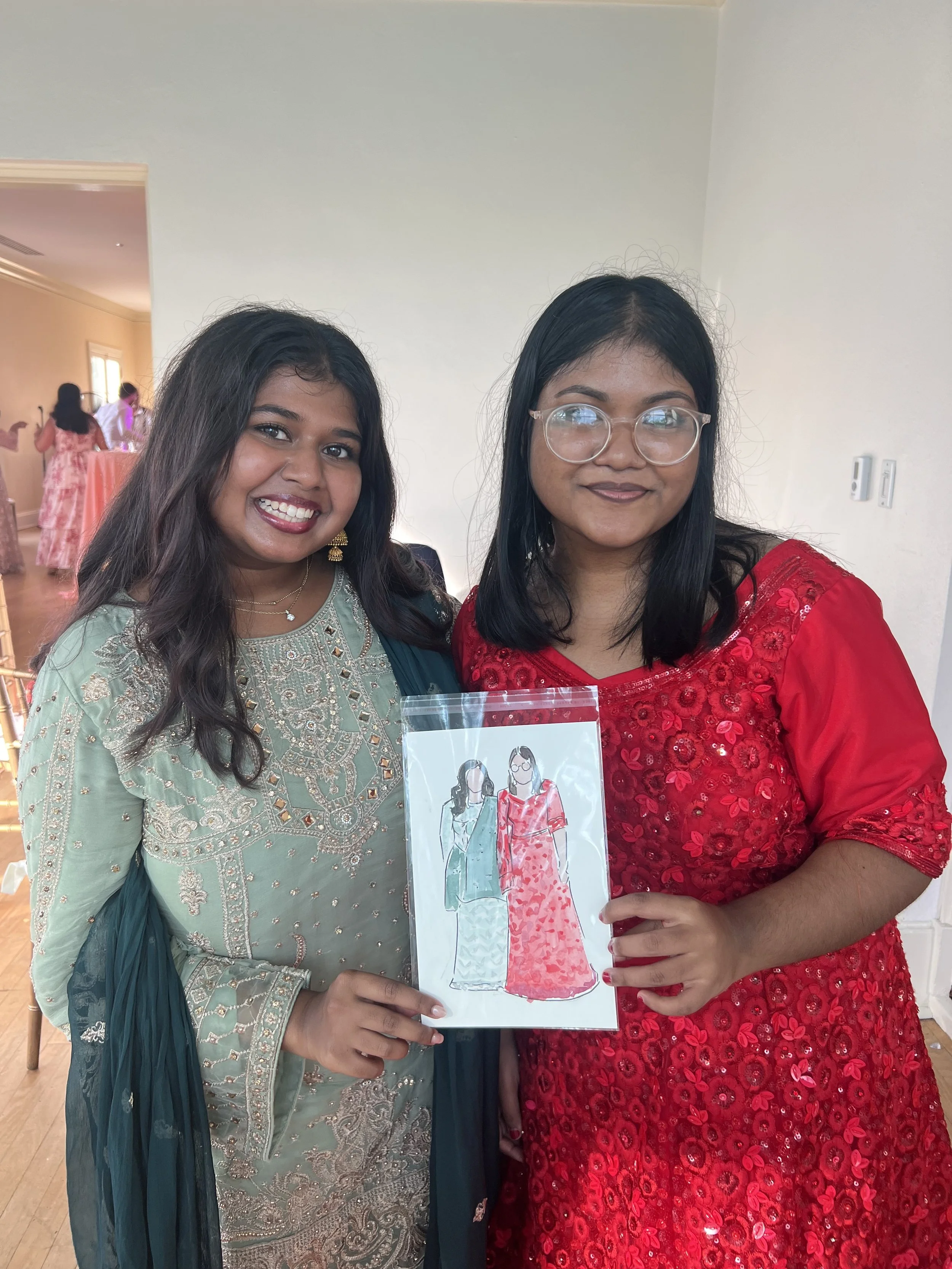 Two women dressed in traditional South Asian attire smiling at the camera while holding a plastic bag with a drawing of two women in festive dresses.