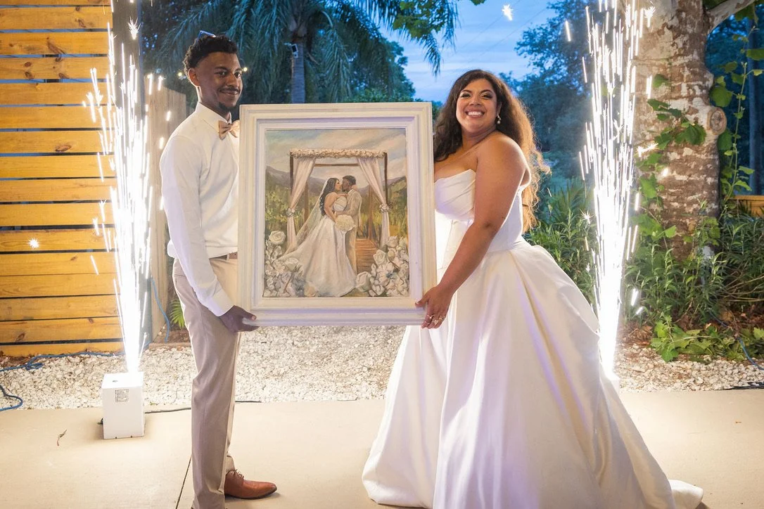 Two people, a man in a white shirt and beige pants and a woman in a white wedding dress, holding a framed painting of a wedding scene during a celebration with fireworks and palm trees in the background.