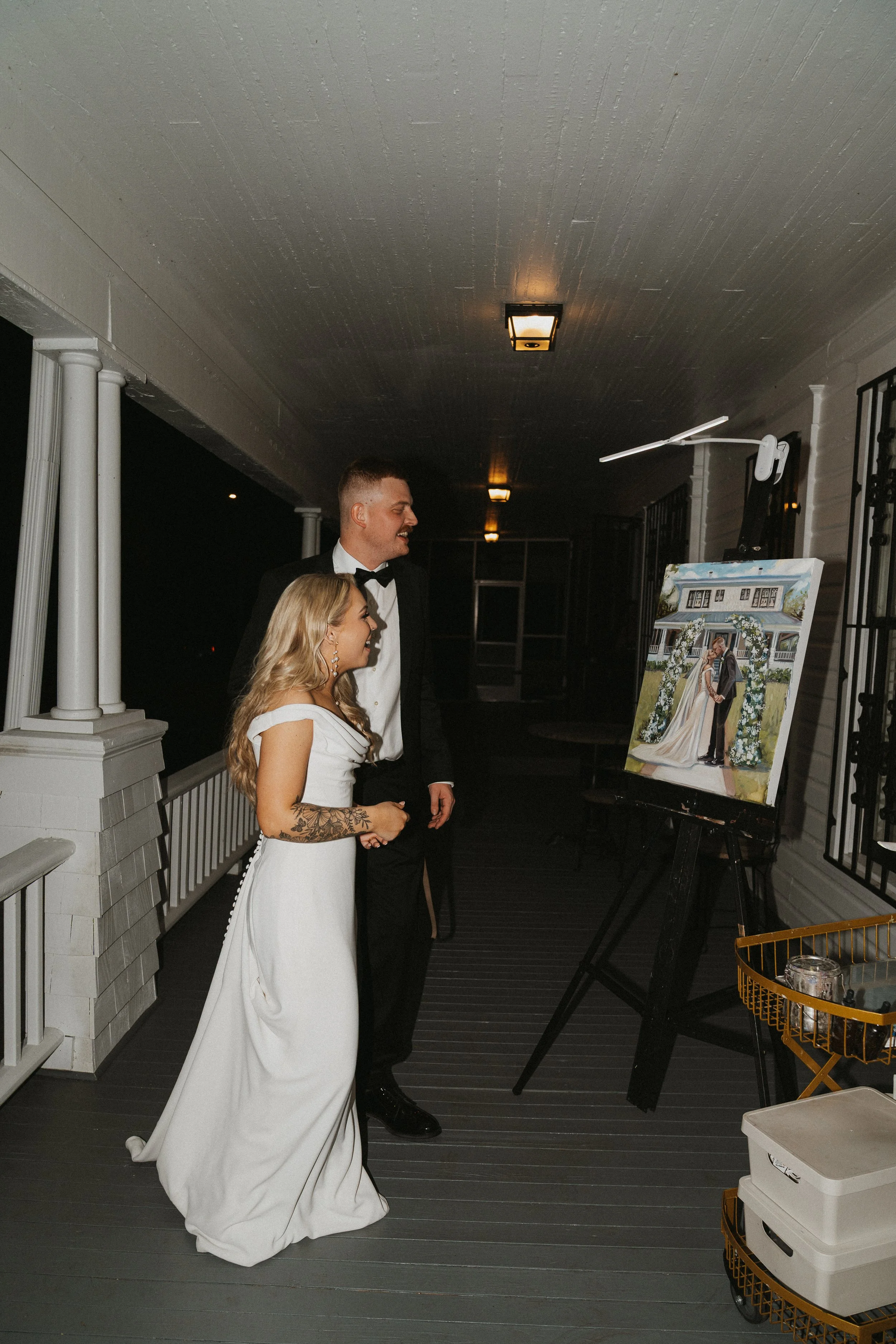 A bride and groom in wedding attire looking at a painting of themselves during their wedding reception on a porch.