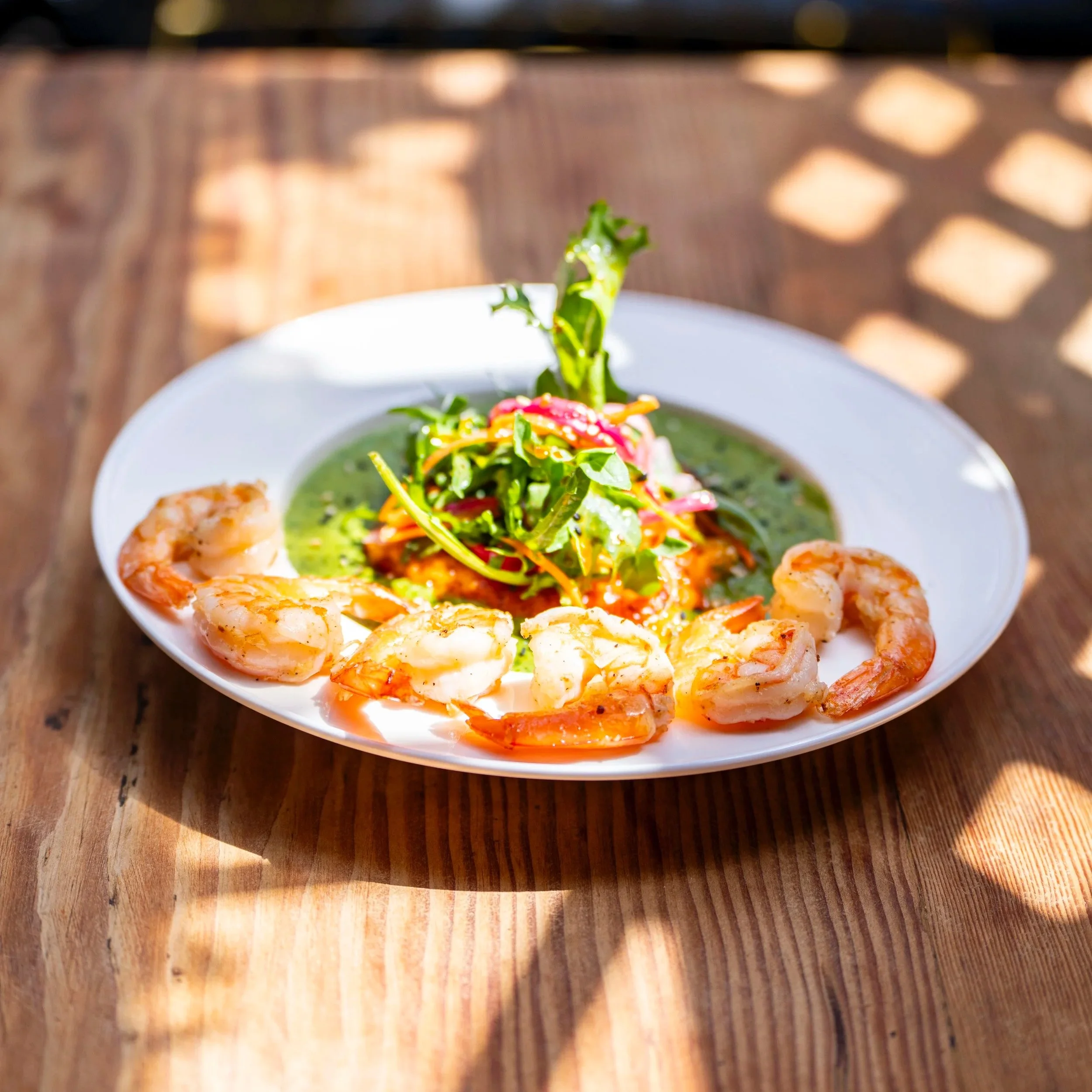 Shrimp salad on a white plate with a green dressing and mixed greens, set on a wooden table.