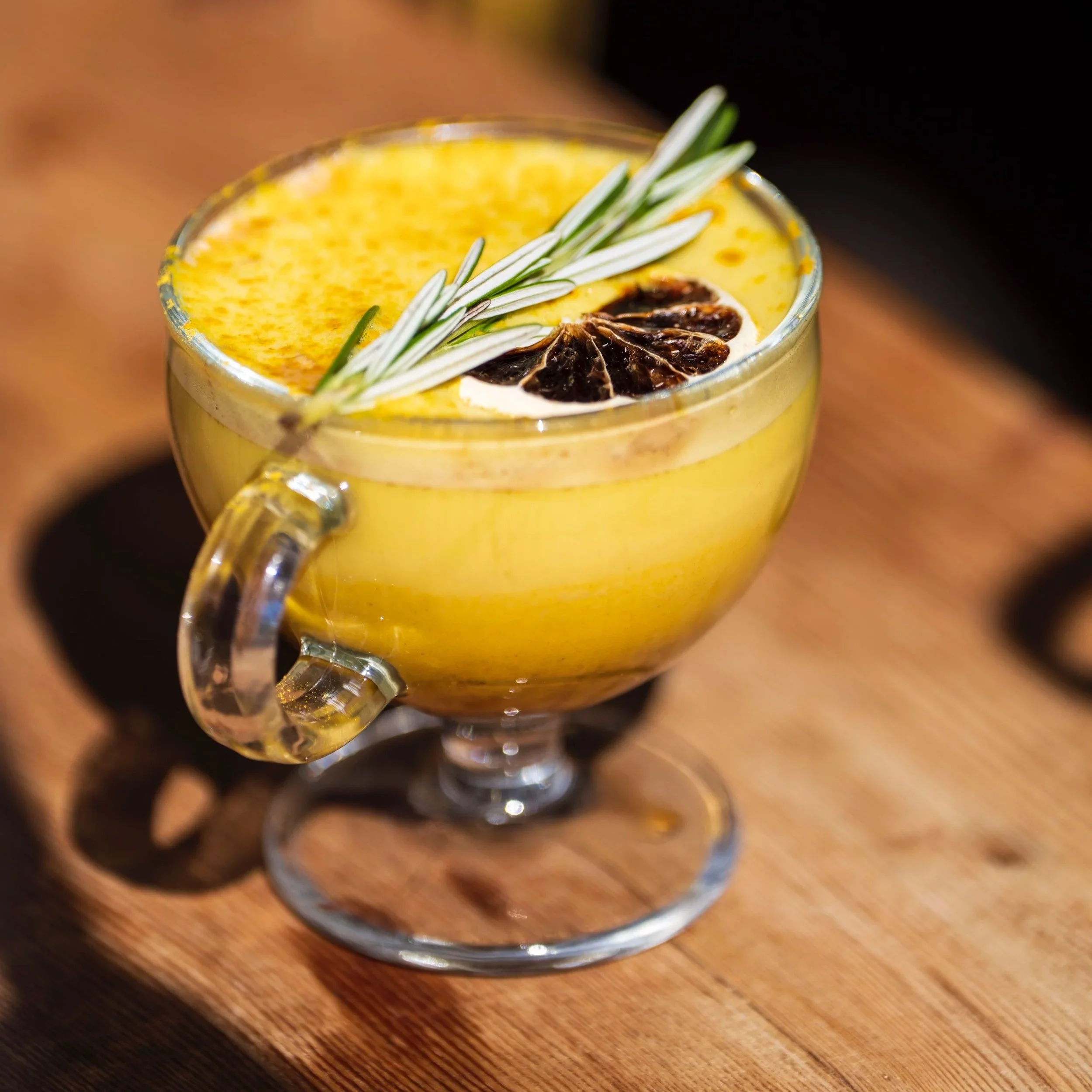 A yellow cocktail garnished with rosemary, a slice of dried lemon, and a dried lime on top, served in a clear glass cup on a wooden surface.