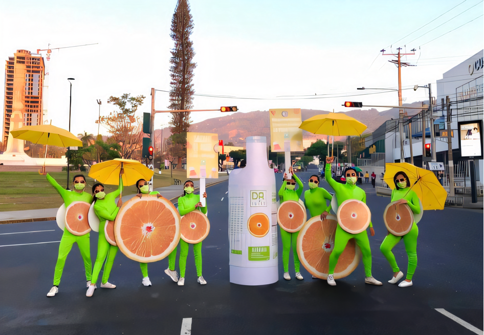 Grupo de personas vestidas de verde lima con diseños de naranja, sosteniendo sombrillas amarillas en una calle con semáforos y edificios, manifestando con una gran botella rotulada "DR. Juice". Activación urbana organizada por Marketing Experience.