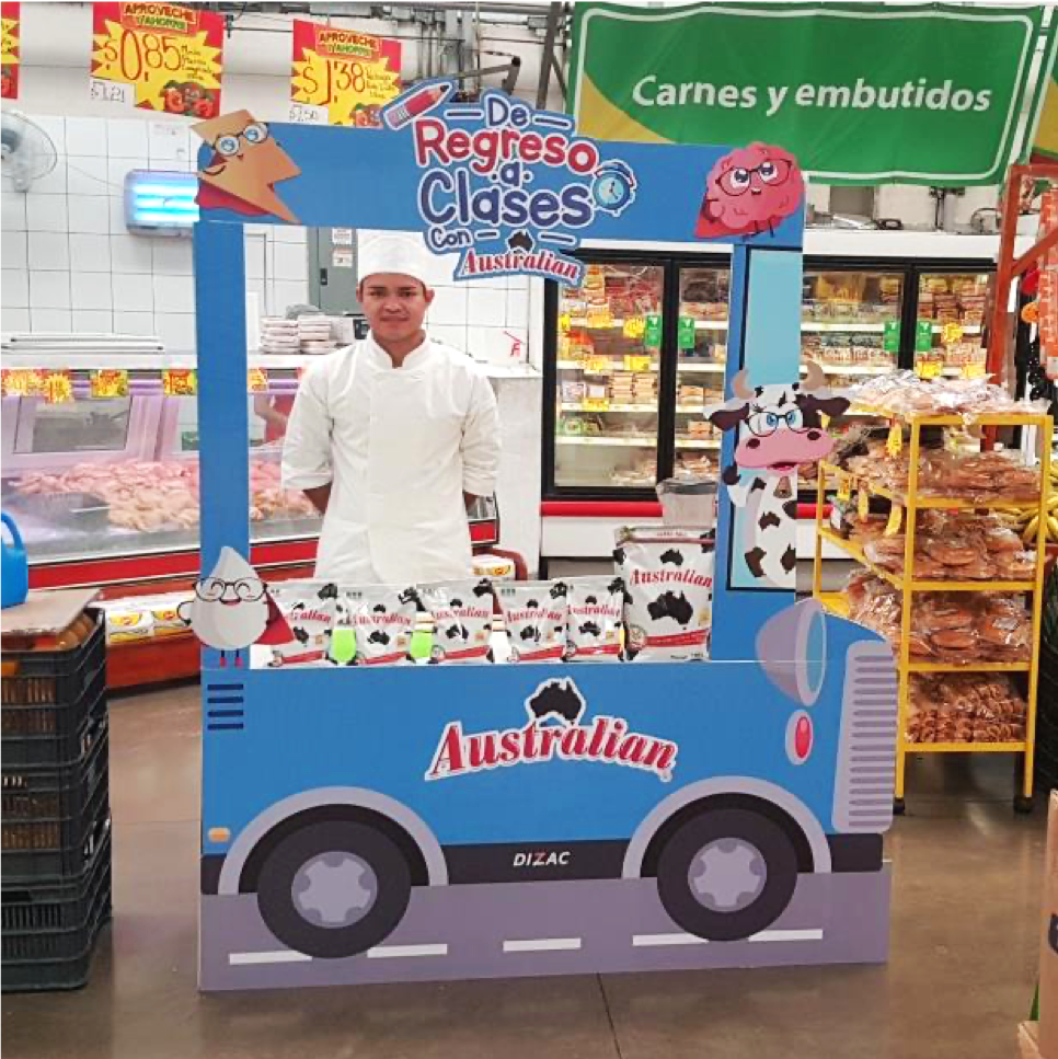 Chef detrás de un stand promocional en un supermercado, decorado con vacas caricaturescas y anuncios de productos Australian. Al fondo se observan áreas refrigeradas de carnes y exhibidores de pan. Activación de Marketing Experience. 