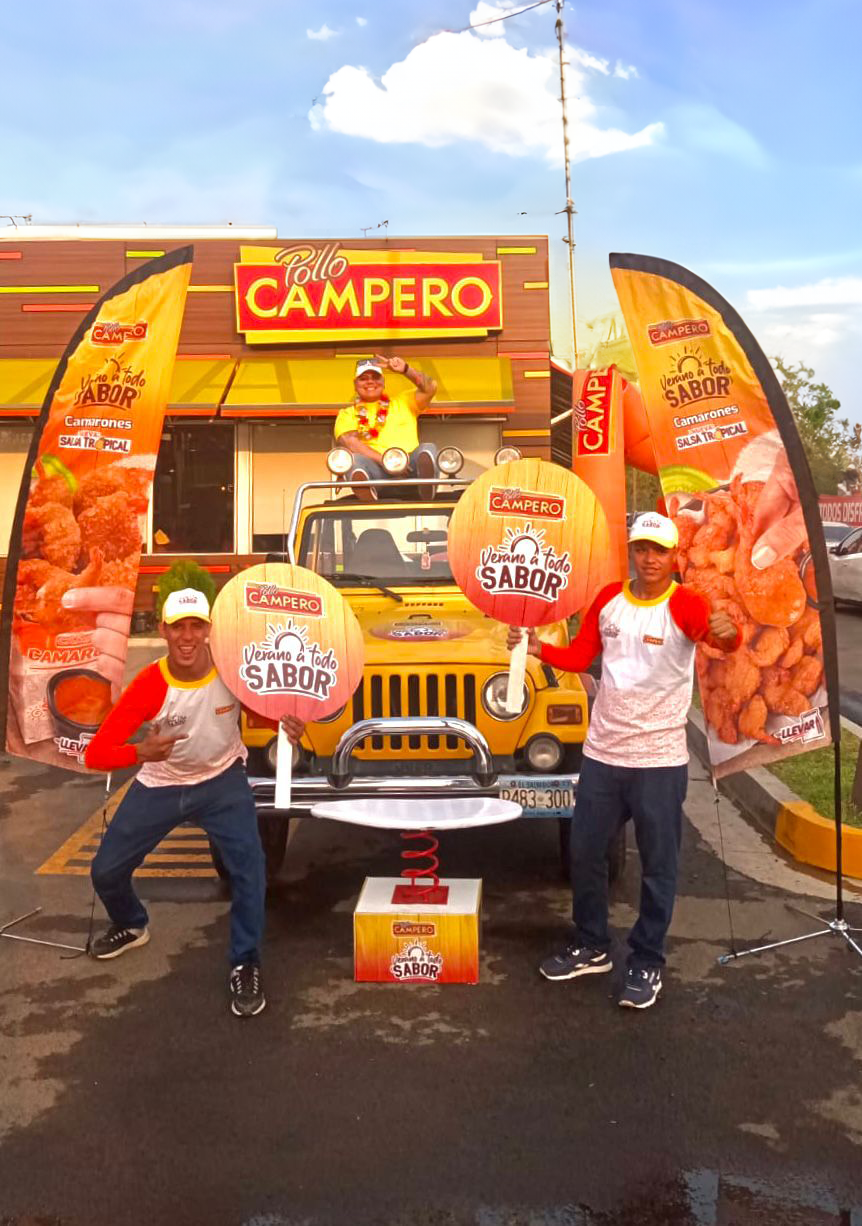 Dos hombres con camisas y gorras rojas y blancas sostienen rótulos redondos con el logo de Campero, frente a un vehículo amarillo decorado con banners de Pollo Campero. Activación organizada por Marketing Experience