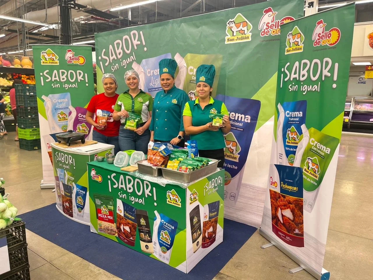 Cuatro mujeres en un stand promocional de San Julián en supermercado, con banners que muestran empaques e imágenes de productos lácteos y el lema “¡SABOR! sin igual”.  Activación organizada por Marketing Experience. 