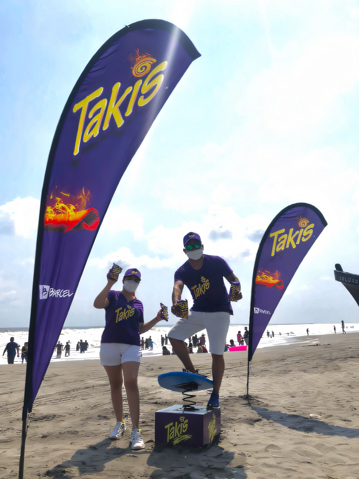 Dos personas sobre una tabla de surf doble con resorte en la arena, sosteniendo paquetes de snacks, rodeadas de banderas moradas con el logo de Takis y un gráfico de snack en llamas. Activación de playa organizada por Marketing Experience. 