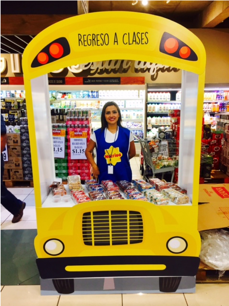 Mujer en supermercado detrás de un recorte en forma de bus escolar amarillo con el letrero “Regreso a Clases”. Sonríe mientras al fondo se ven estantes con productos. Activación organizada por Marketing Experience. 