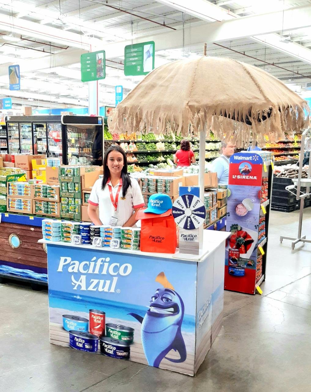 Una mujer en un stand promocional de Pacífico Azul dentro de un supermercado, con una sombrilla grande tipo playera y latas del producto exhibidas sobre el mostrador. Activación organizada por Marketing Experience.