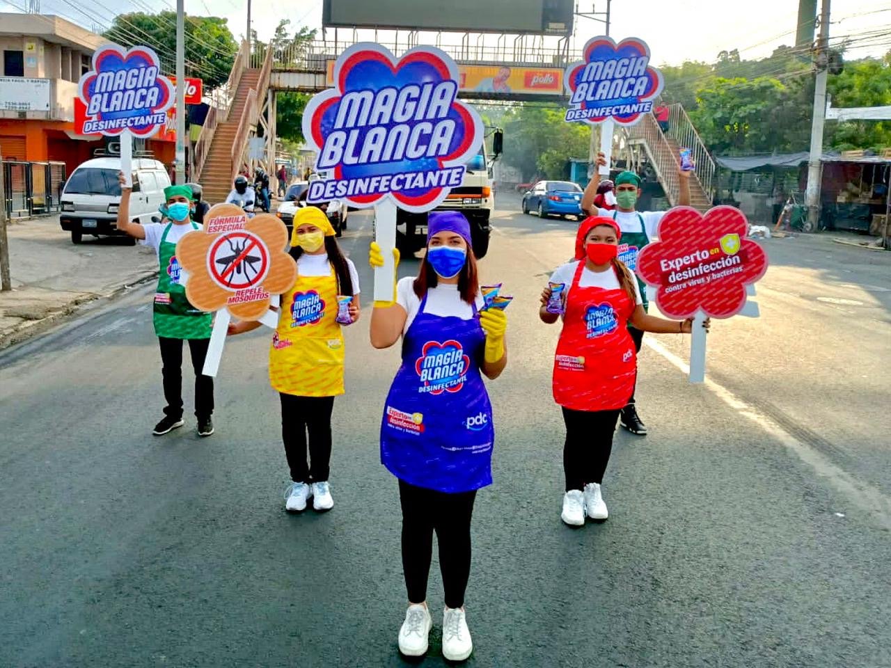 Grupo de personas con delantales coloridos y mascarillas, sosteniendo carteles que promueven el desinfectante Magia Blanca contra los mosquitos, en una calle con carros y edificios de fondo. Activación organizada por Marketing Experience. 