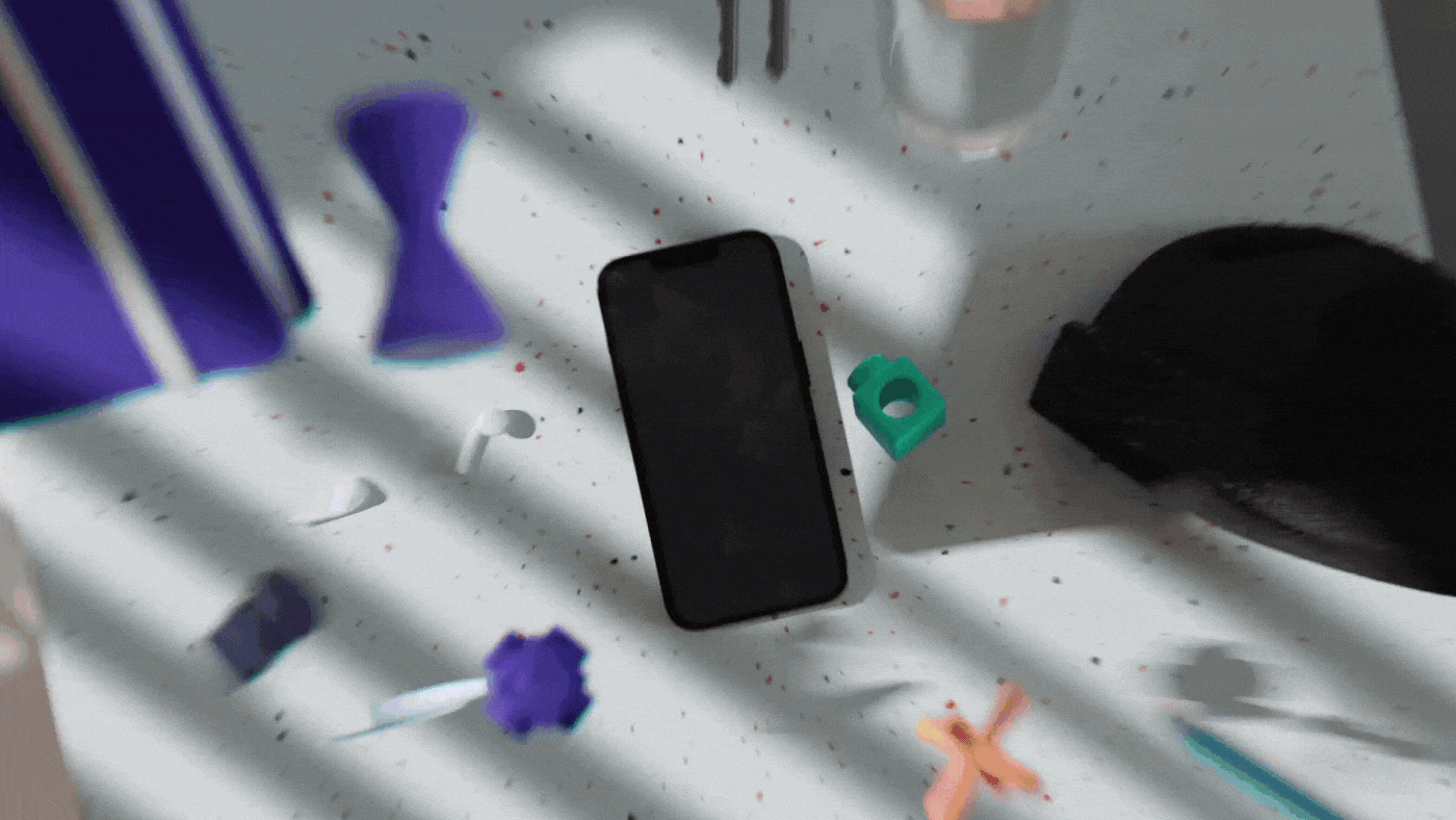 A smartphone lying on a white surface with various small tools and objects around it, including purple and green objects, a black cloth, and colorful splatters.