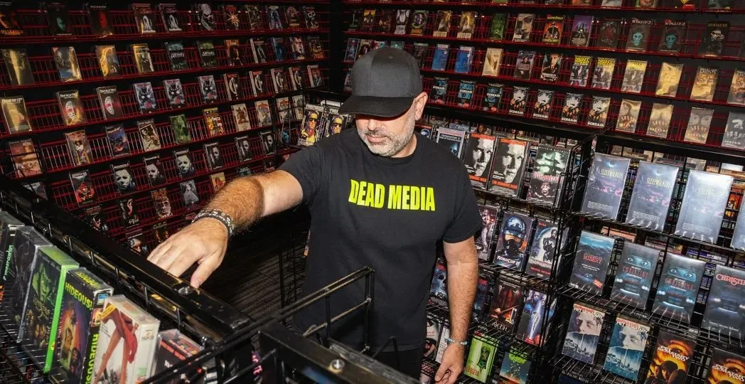 Jonathon Carmichael, founder, is wearing a black shirt that says "Dead Media". He is browsing the racks of the video rental exhibit in the Killer Video museum.