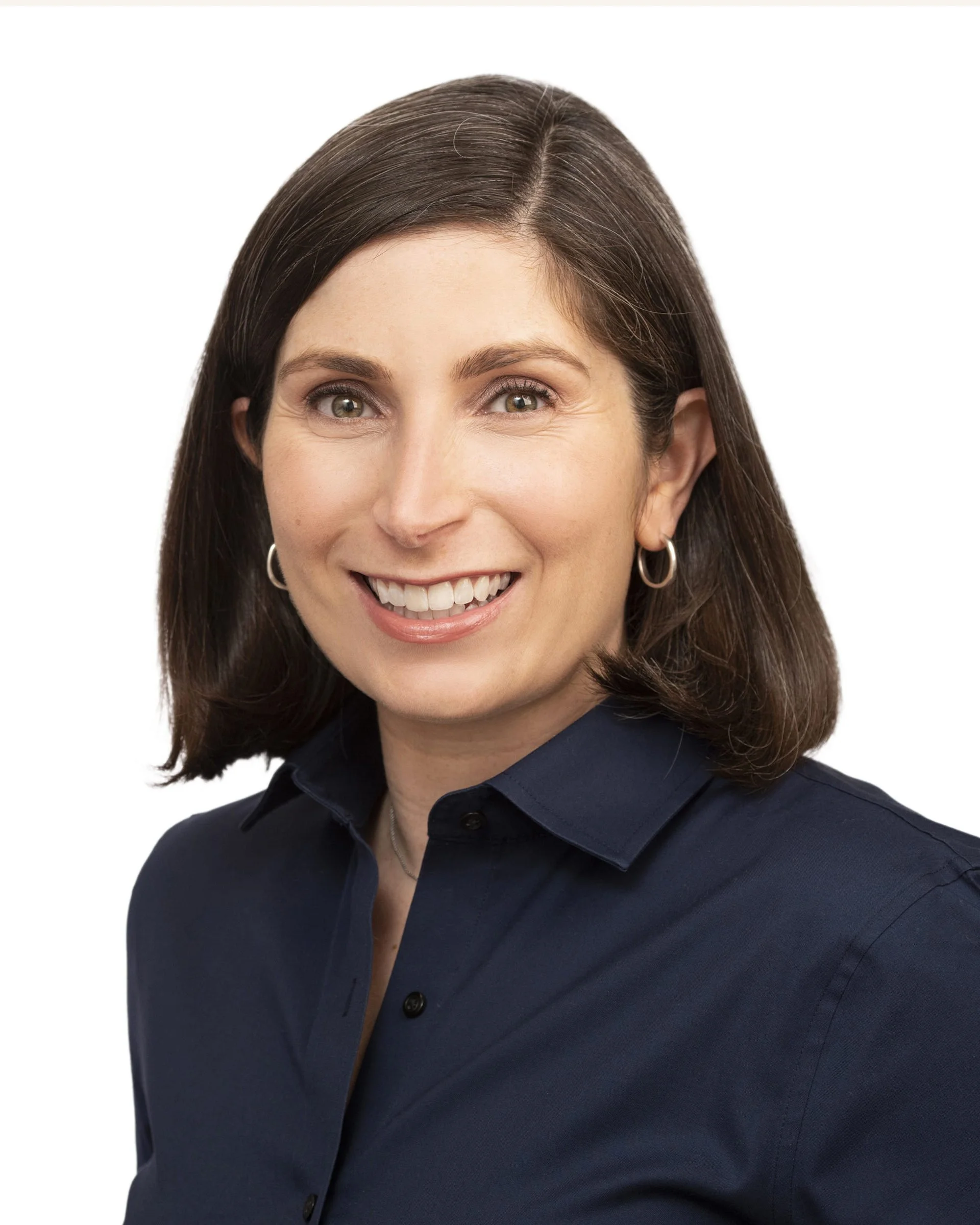 A woman with shoulder-length brown hair, wearing a dark blue button-up shirt and small hoop earrings, smiling against a white background.