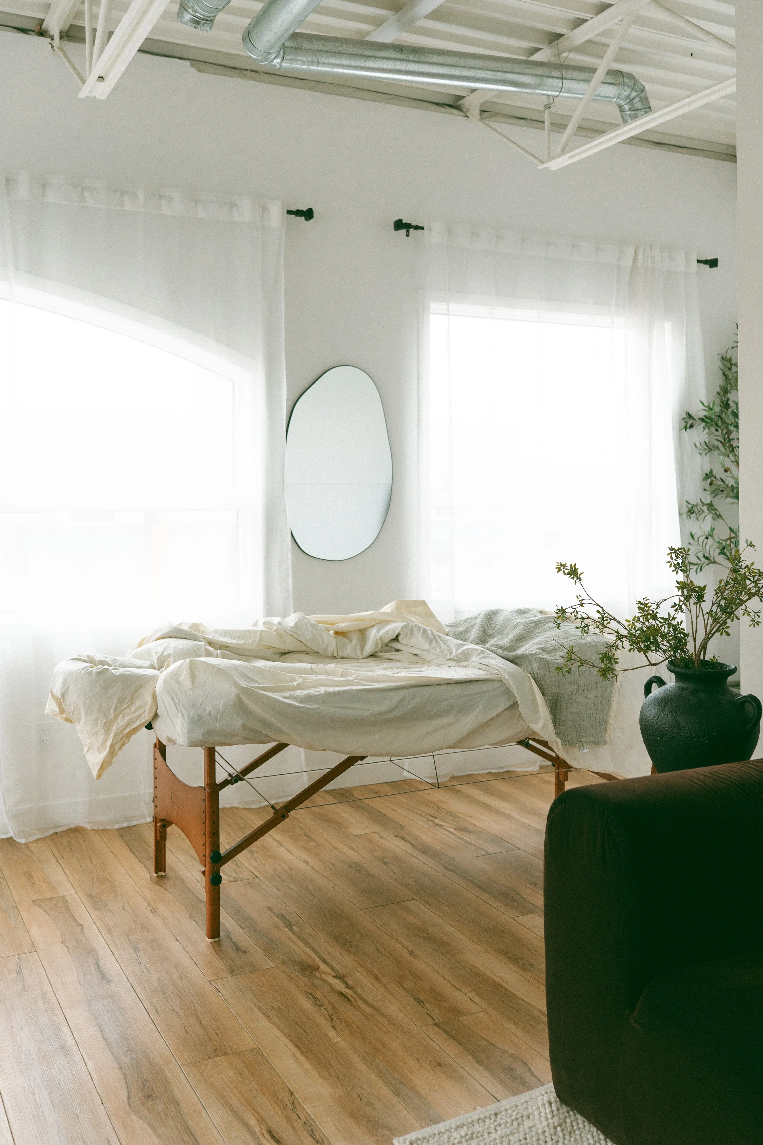 A massage bed with white sheets and a blanket in a bright room with wooden floors, large windows with white curtains, a mirror on the wall, and a large potted plant.
