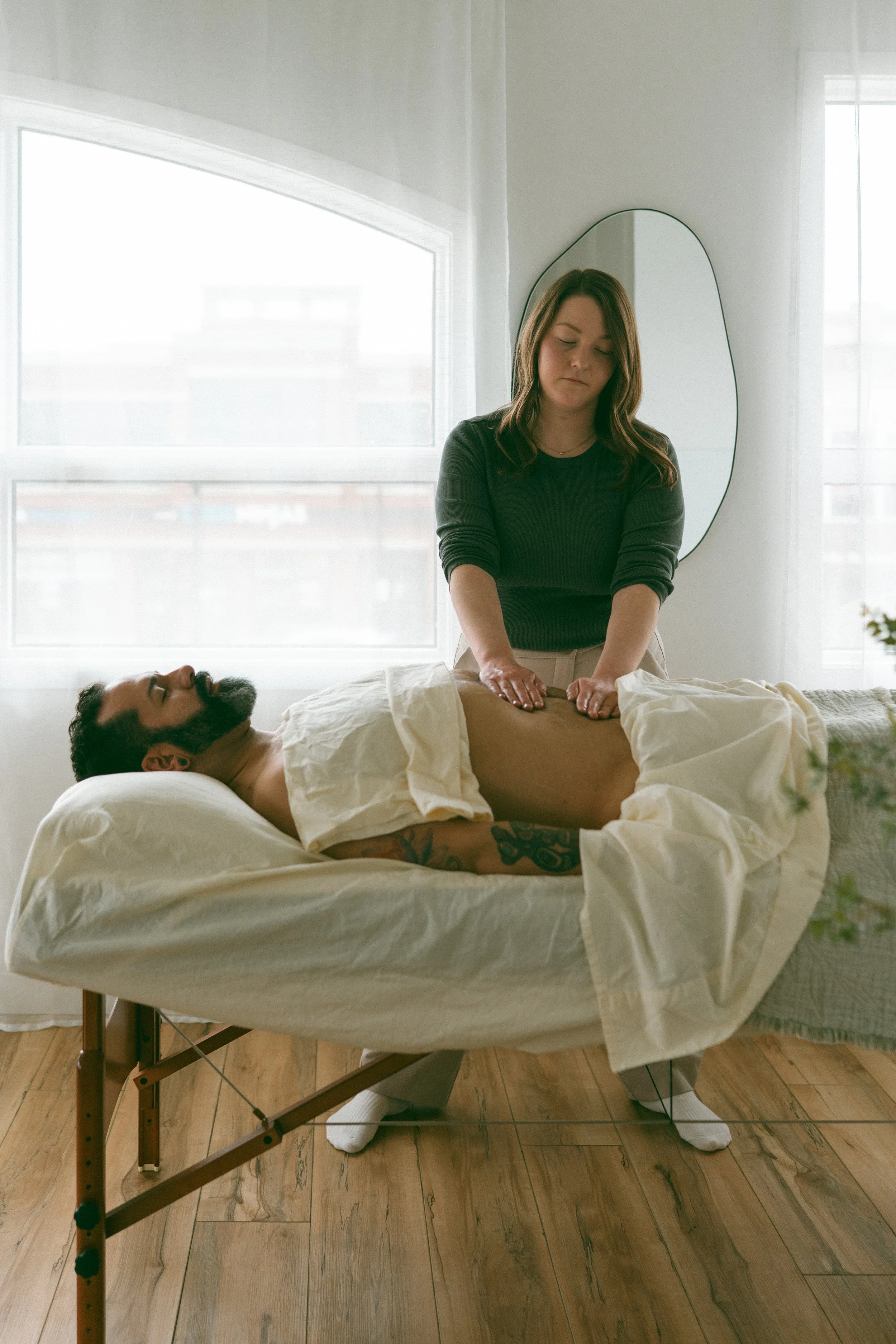 A woman giving a massage to a man lying on a bed. The woman has brown hair and is wearing a dark top, while the man has a beard and tattoos on his arm. The room is bright with large windows and a sleek mirror on the wall.
