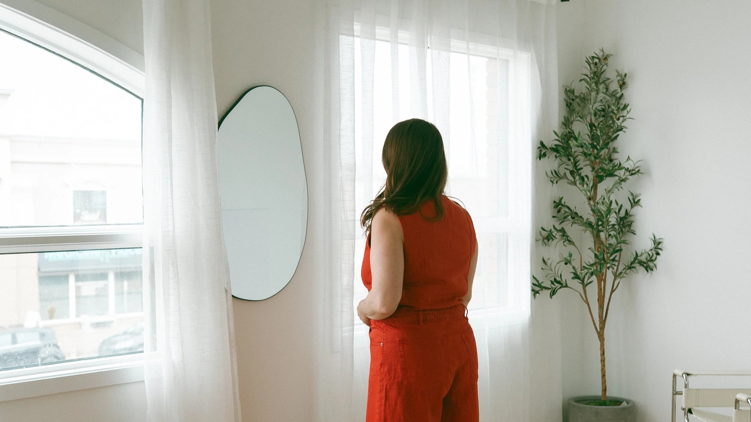 A woman with brown hair wearing a red sleeveless top and red pants stands in a bright room by a window, looking at a mirror. There are white sheer curtains on the window, a potted plant with green leaves on the right, and part of a white chair in the lower right corner.