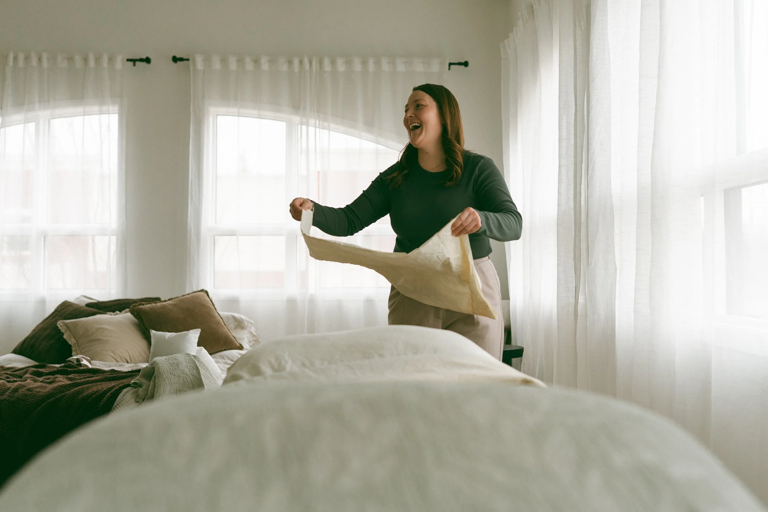 Woman standing by a bed, laughing, holding a pillow, in a bright room with large windows and sheer curtains.