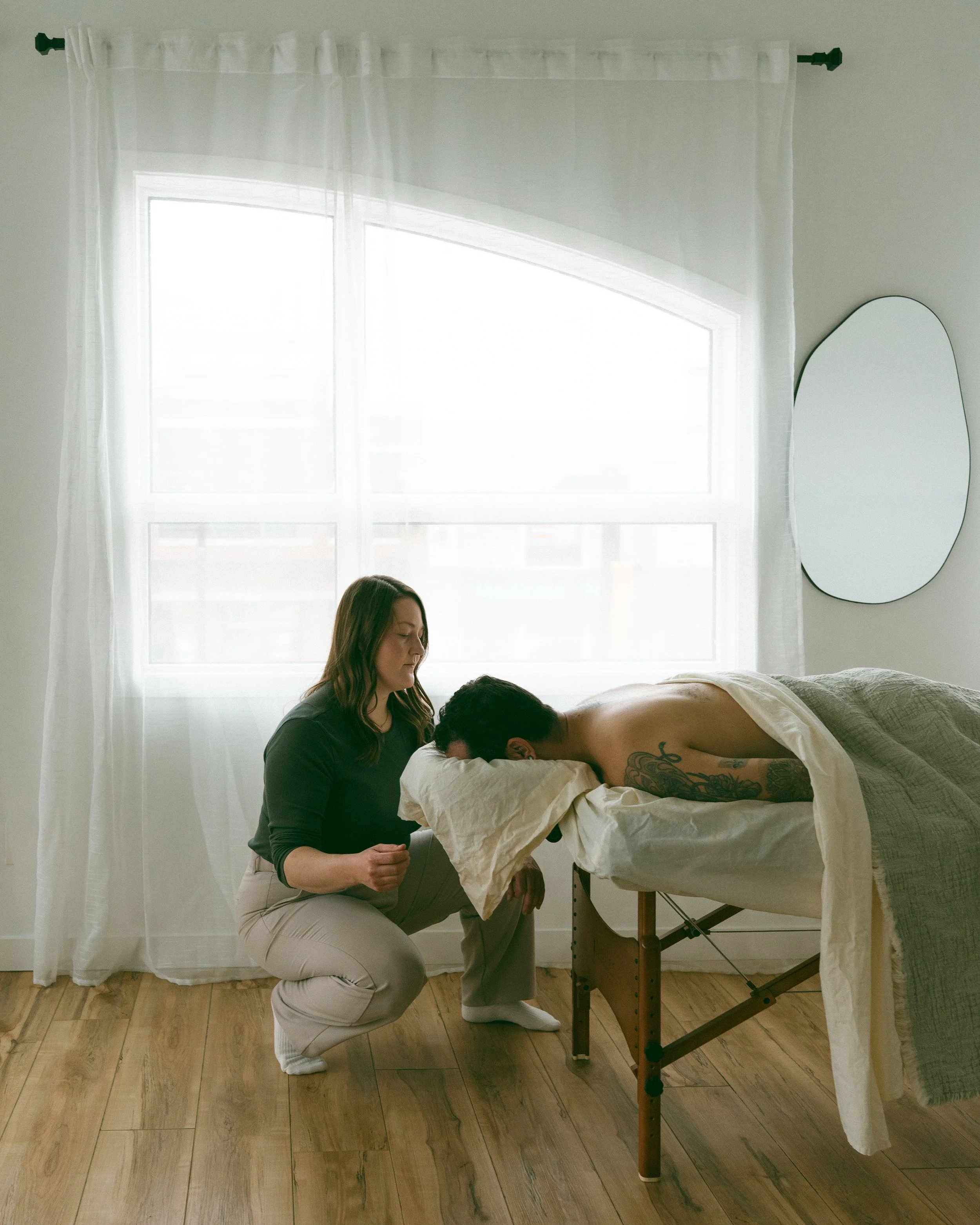 A woman kneeling beside a massage table, giving a massage to a person lying face down on the table, with a white curtain and window in the background.