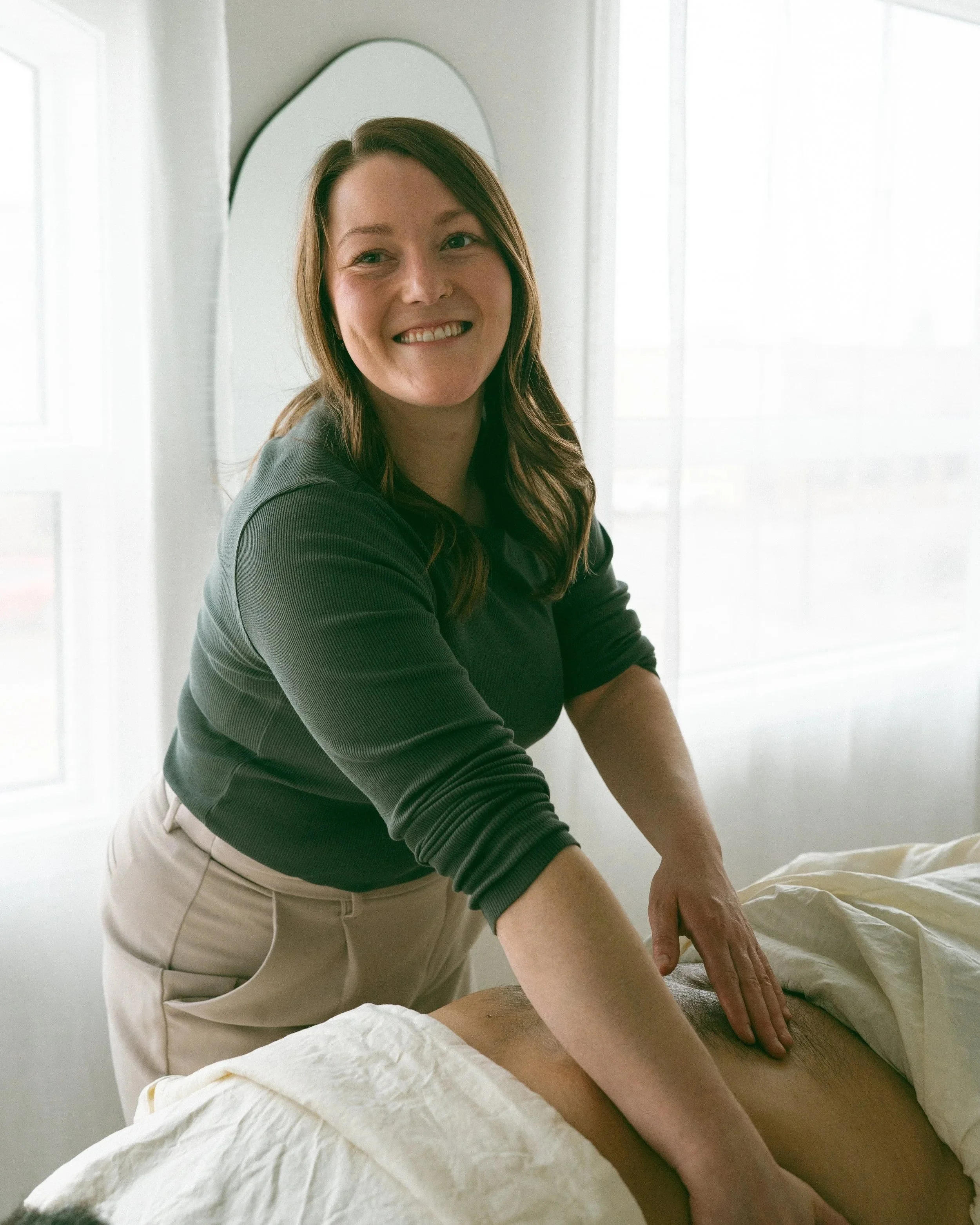 Emma giving a back massage to a man lying face down on a massage table, in a bright room with white walls and curtains.