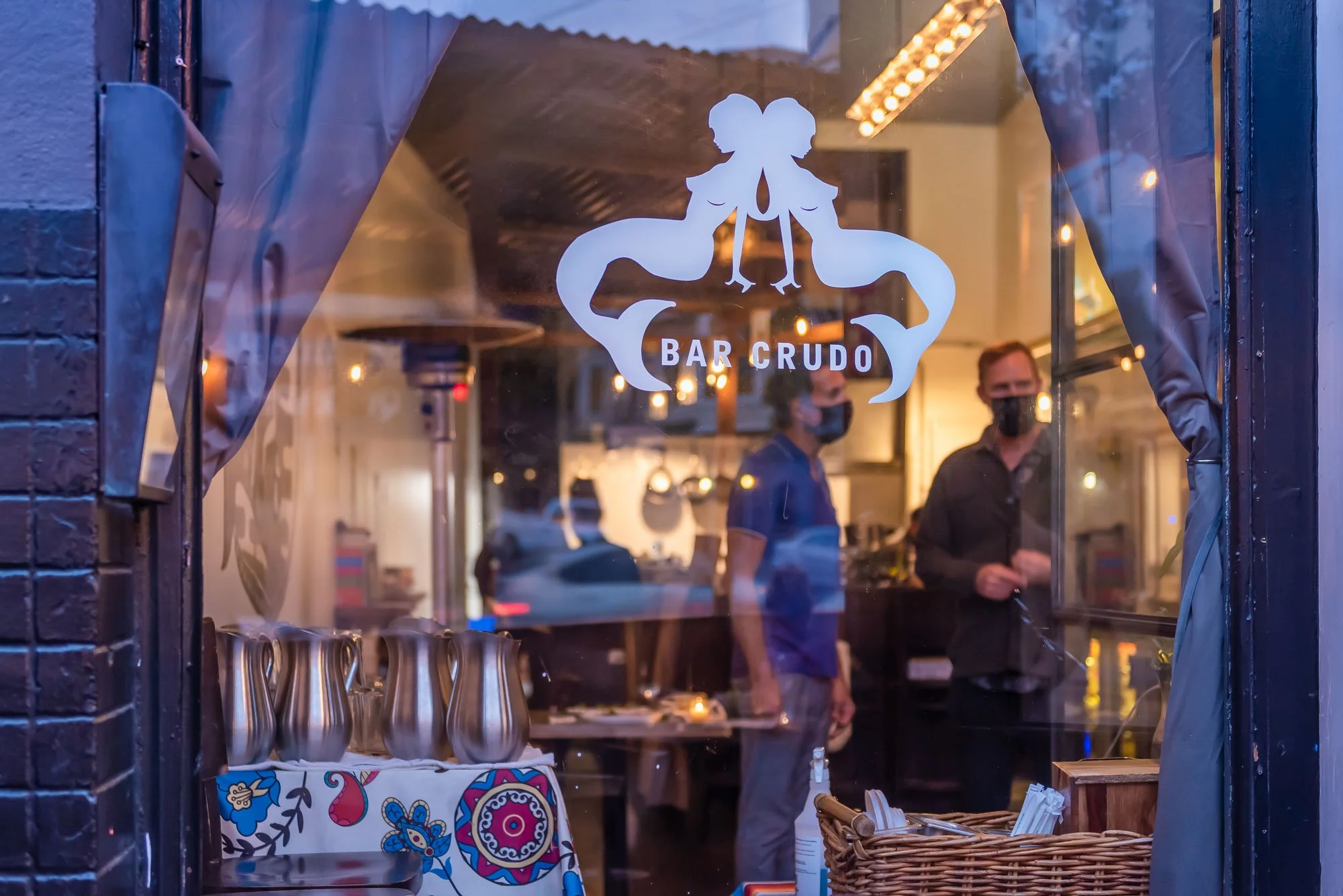 View through the window of Bar Crudo, with people inside wearing face masks and a display of metal pitcher jugs and a basket of utensils on the window sill.
