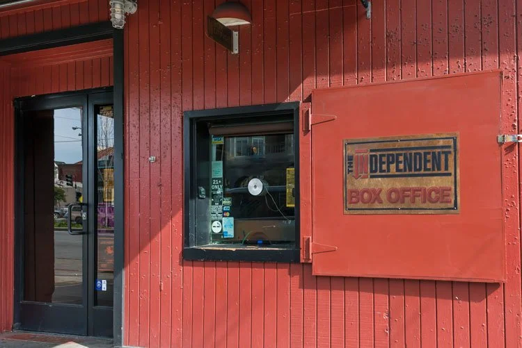 Red exterior wall of a building with a black door, a window with a white speaker, and a sign that reads "The Independent Box Office" with a red background and black text.