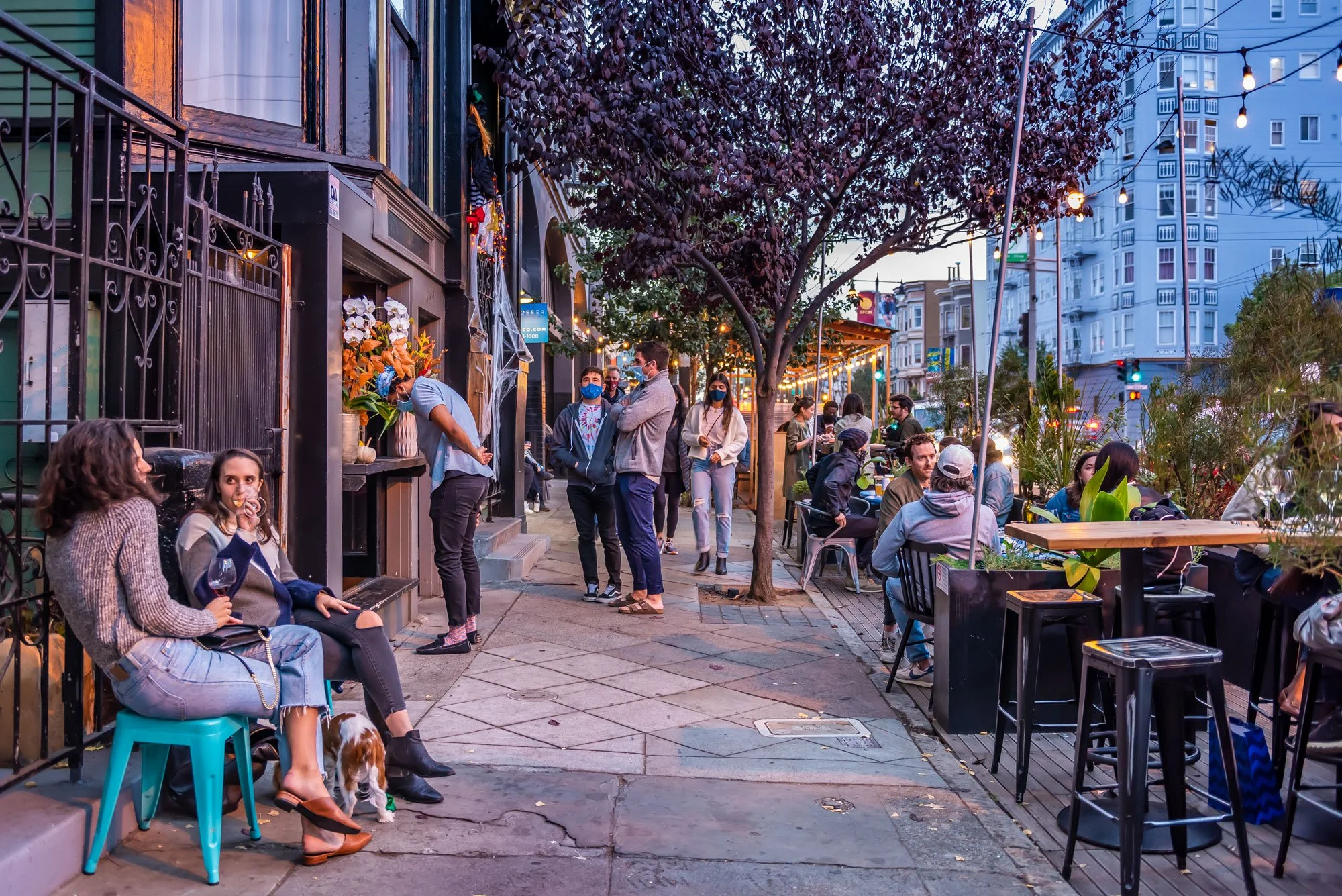 Urban outdoor dining scene at dusk with people sitting at tables, socializing, and walking on the sidewalk, decorated with string lights and a central tree